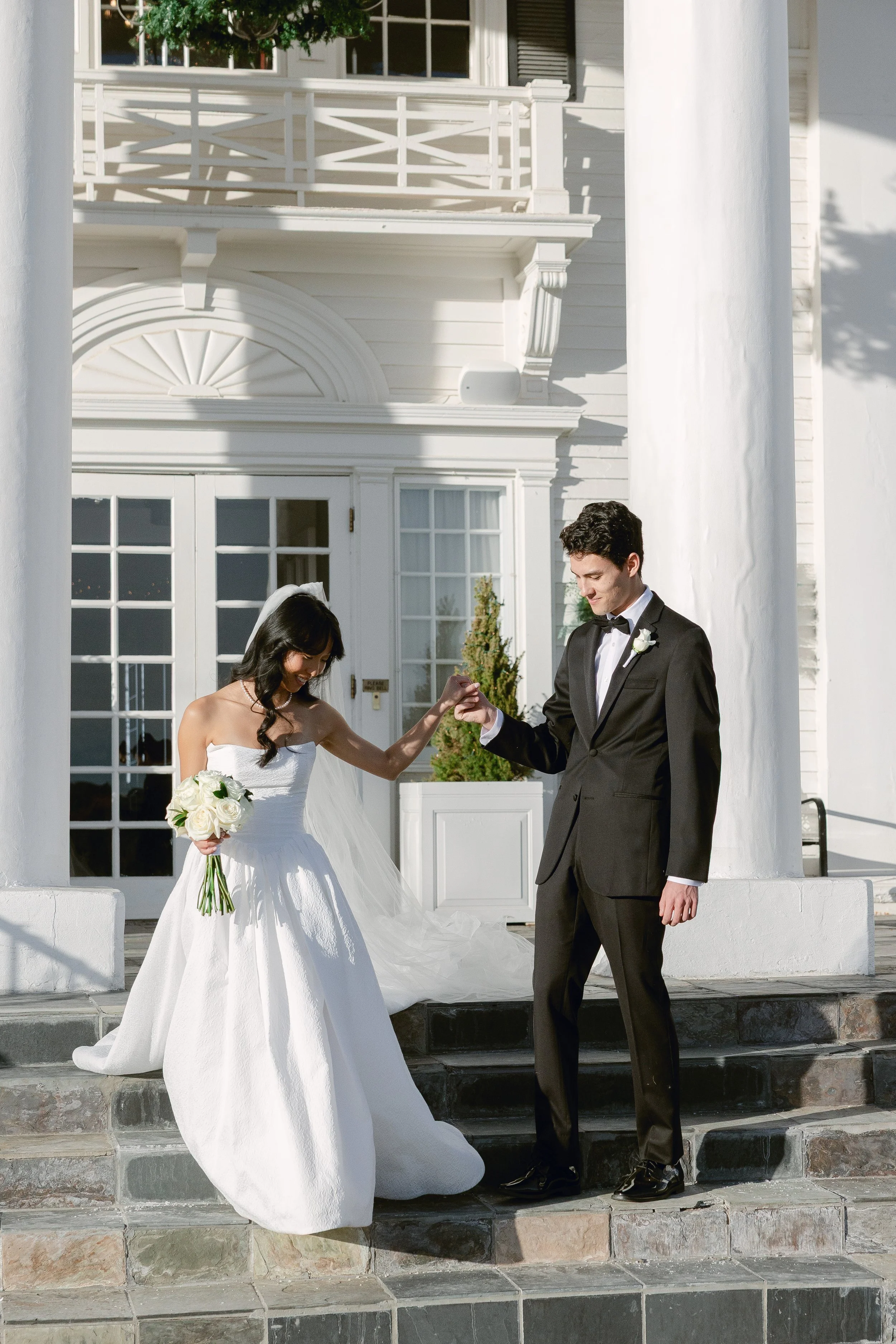 A bride and groom standing on steps outside a white building, holding hands and smiling. The bride is holding a bouquet of white roses and wearing a white wedding dress, and the groom is in a black tuxedo.
