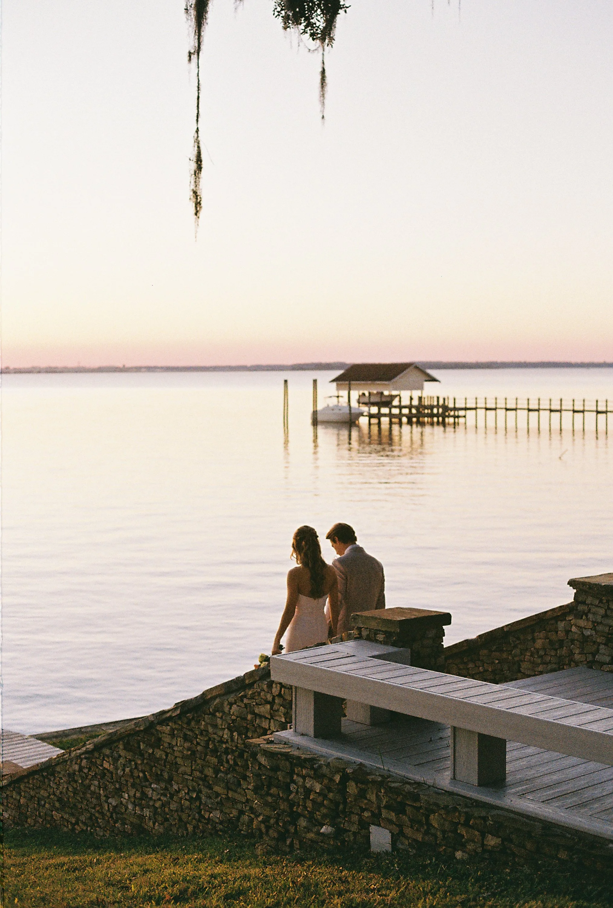A couple dressed in wedding attire standing on stone stairs by the water at sunset, with a dock and boat house in the background.