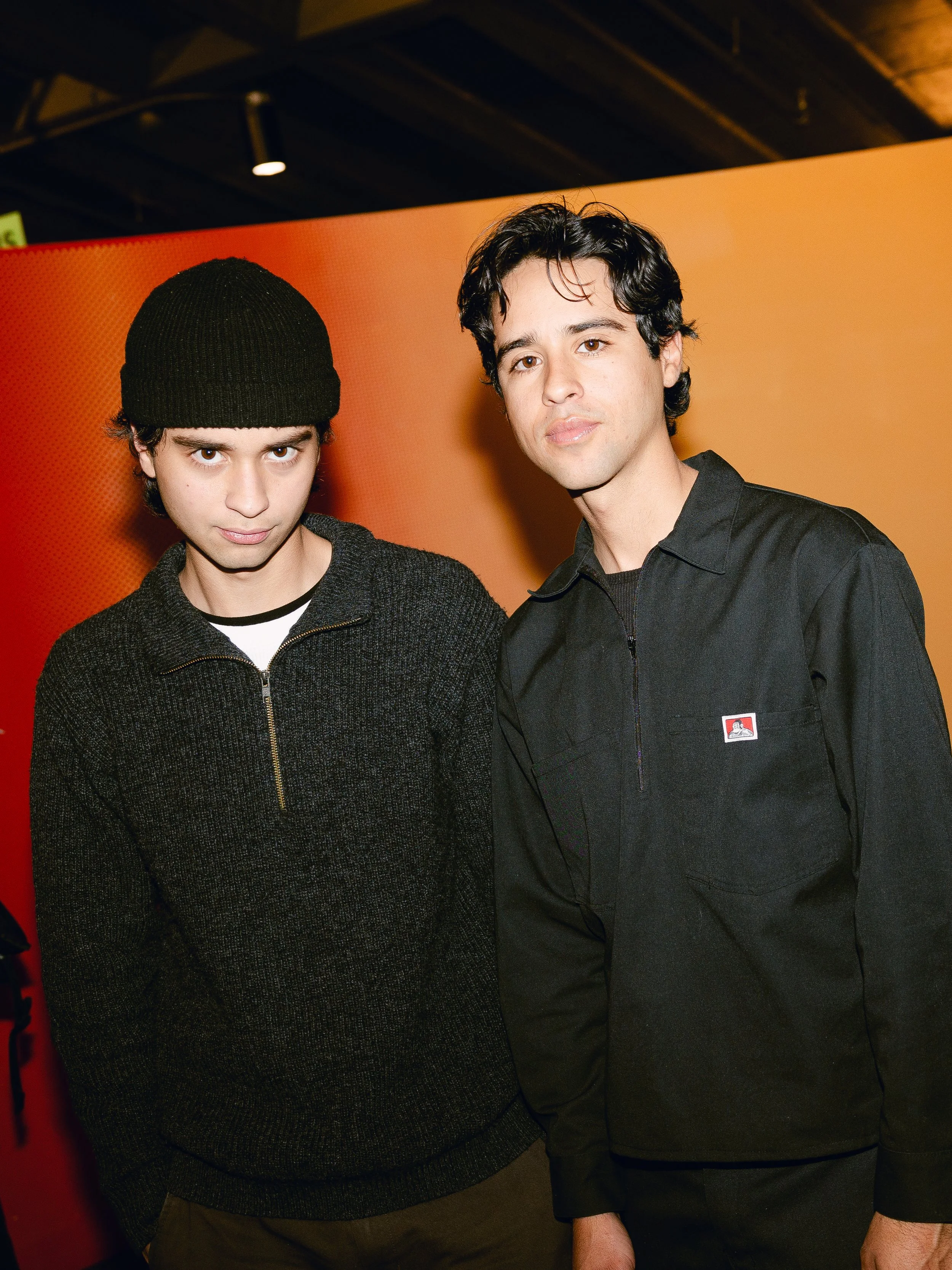 New York City Movie Premiere. Two young men standing side by side in front of an orange background, one wearing a black beanie and dark sweater, the other with dark hair wearing a black button-up shirt with a small patch on the chest.