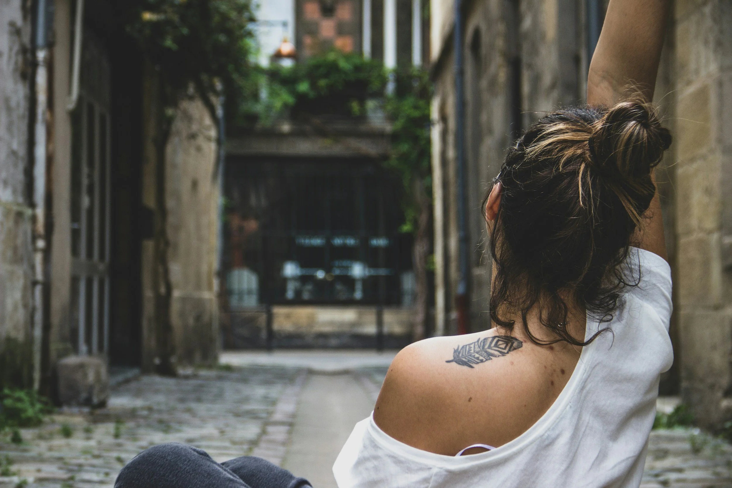 Back view of a person with a bun hairstyle sitting in a narrow cobblestone alley, wearing a white top, showing a feather tattoo on their shoulder.