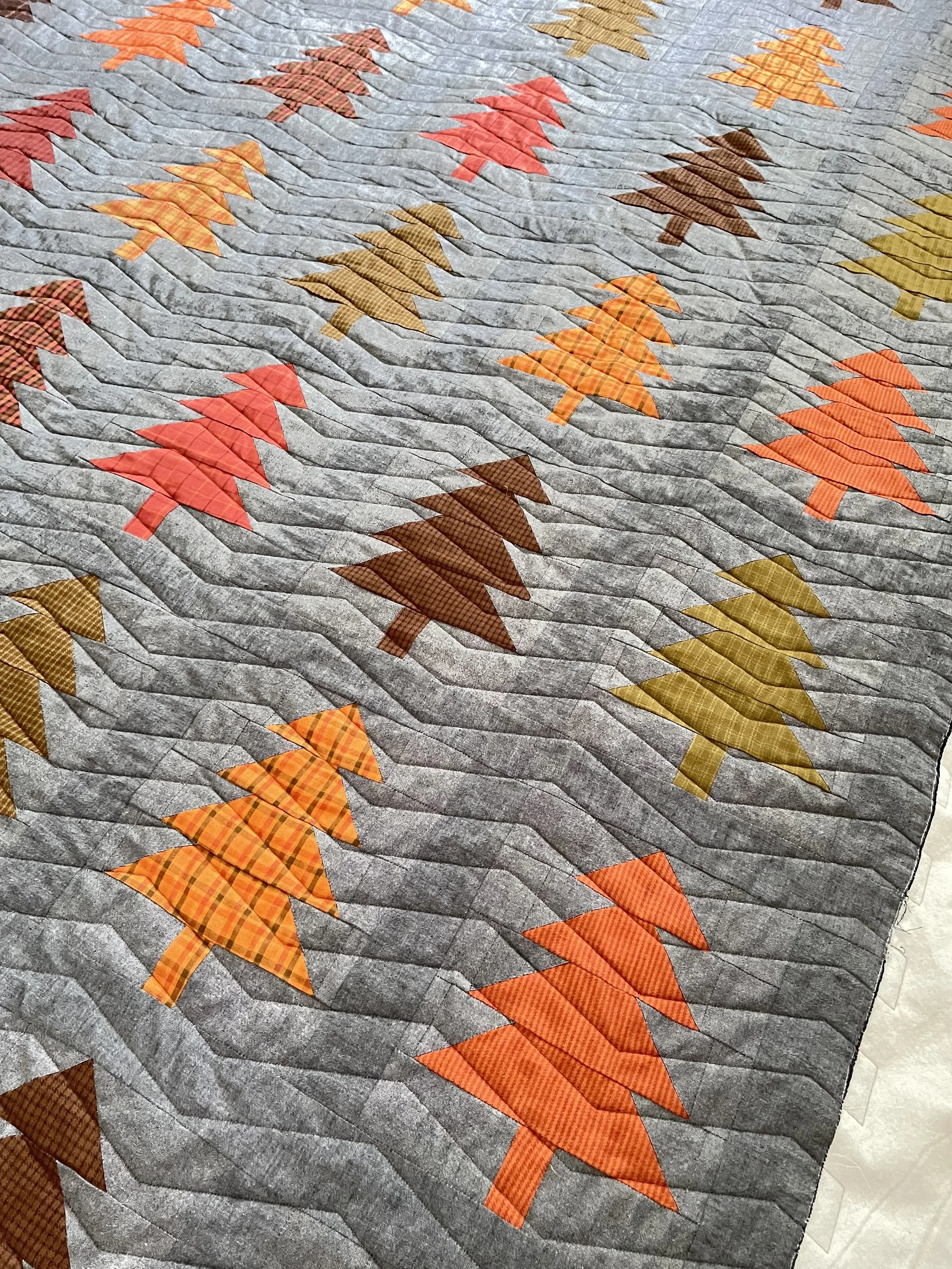 Close-up of a quilt with a grey background and a pattern of colorful triangular trees in shades of orange, red, brown, and green.
