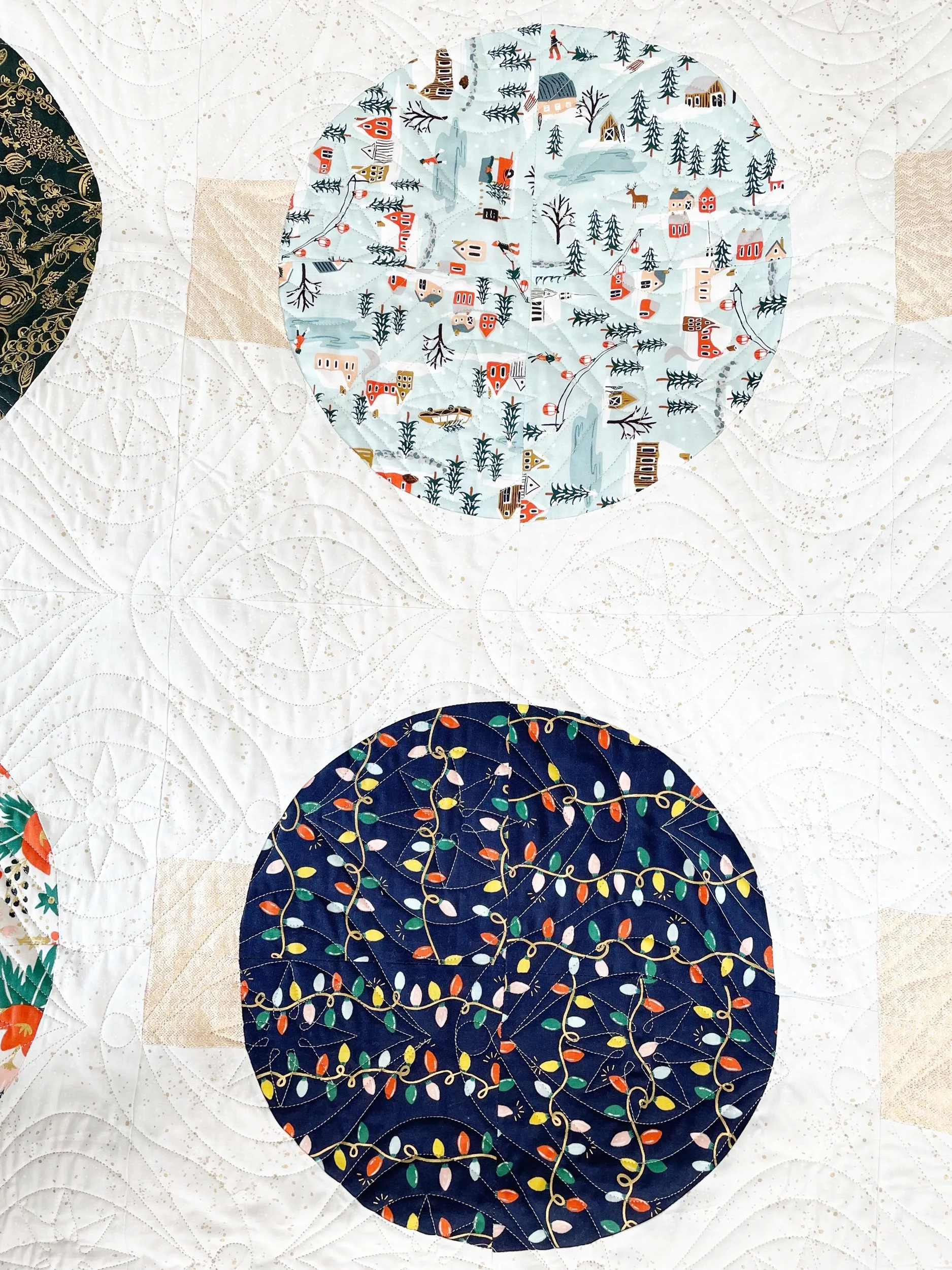 Close-up of a quilt with two large circular fabric patterns, one featuring a winter village scene and the other decorated with Christmas lights.