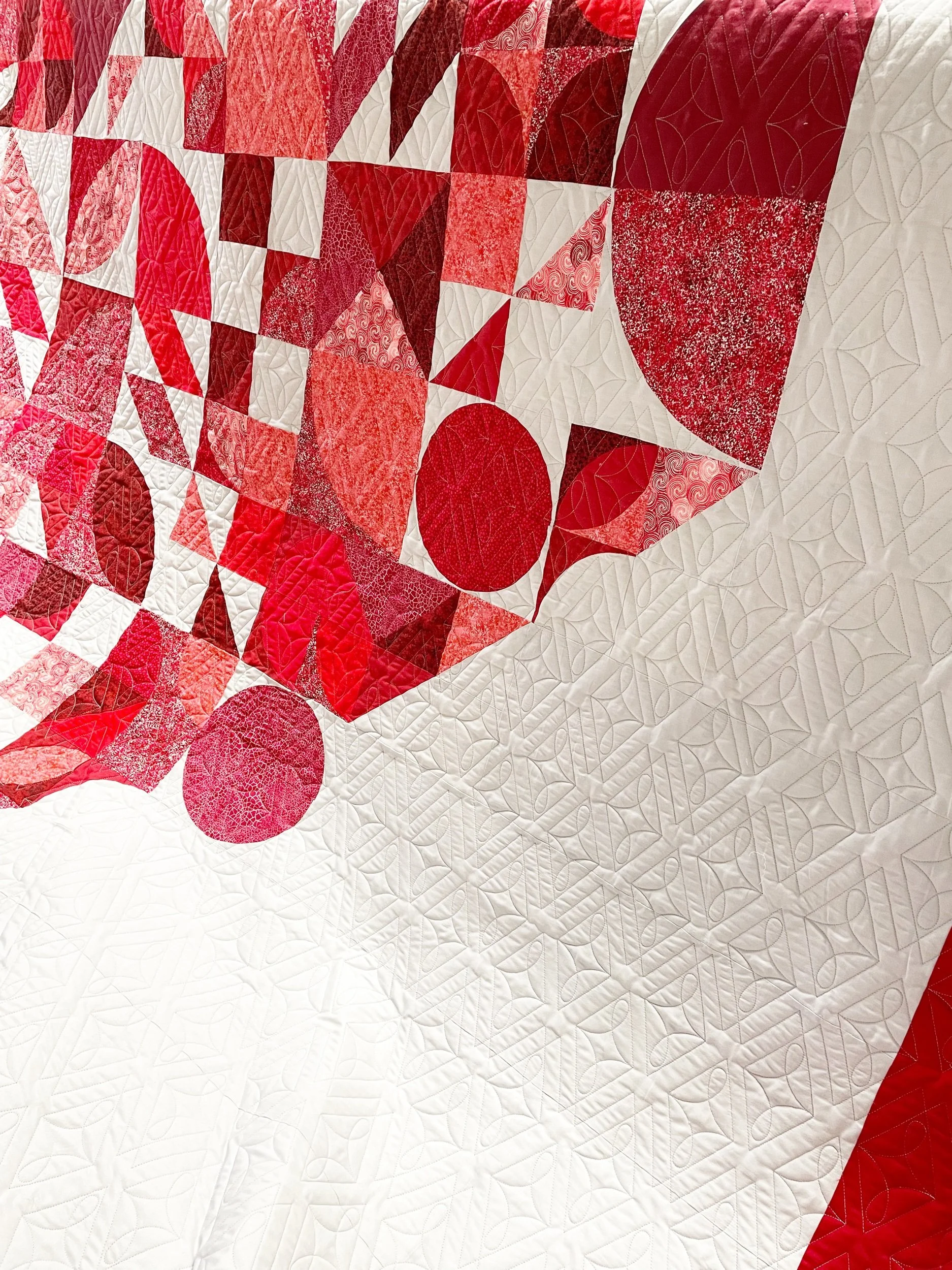 Close-up of a quilt with many red, white, and pink fabric squares, featuring various patterns and designs, including circles, triangles, and abstract shapes.