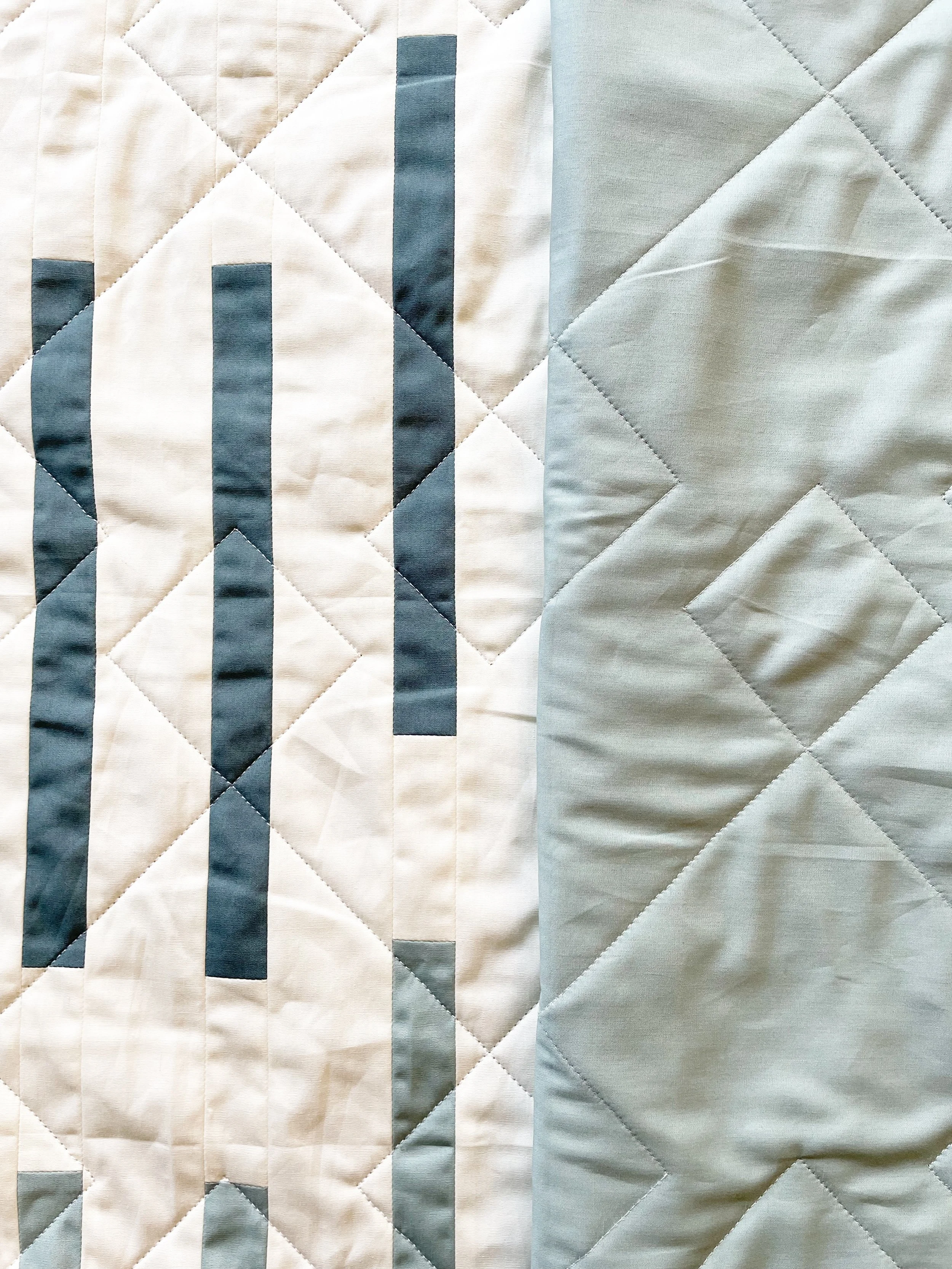 Close-up of a quilt with a geometric pattern, featuring vertical dark blue, light blue, and gray rectangles on a cream background on the left, and a solid light blue section on the right.