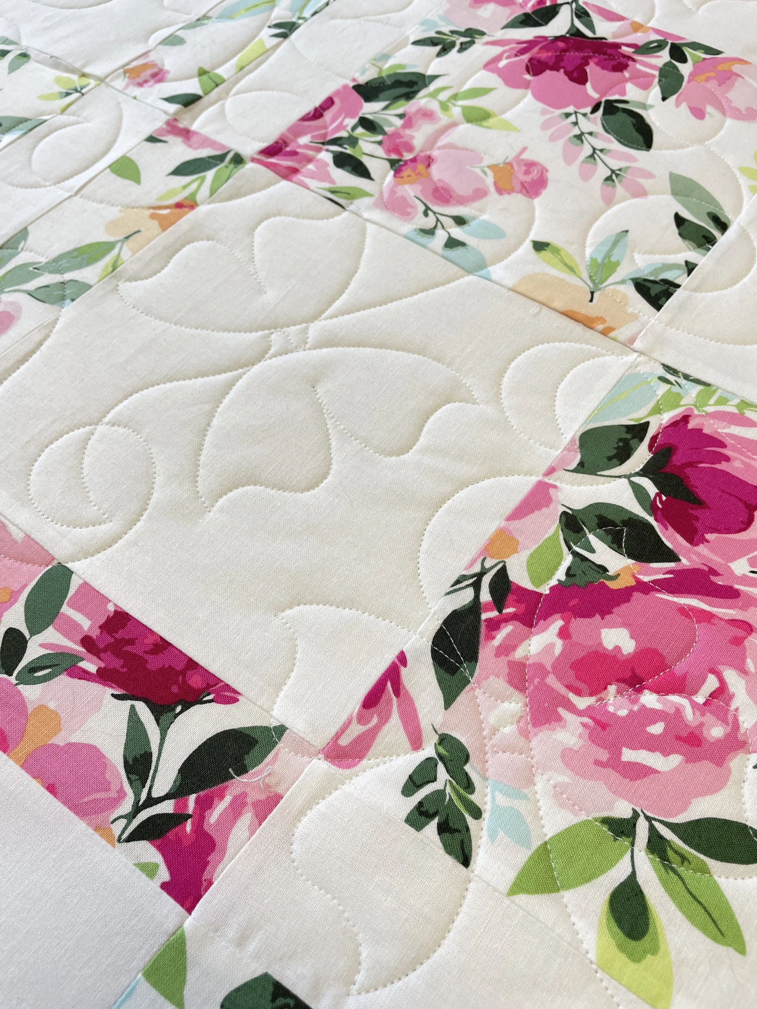 Close-up of a patchwork quilt with floral patterns, featuring cream-colored fabric with stitched outlines and sections of pink, green, and peach floral designs.