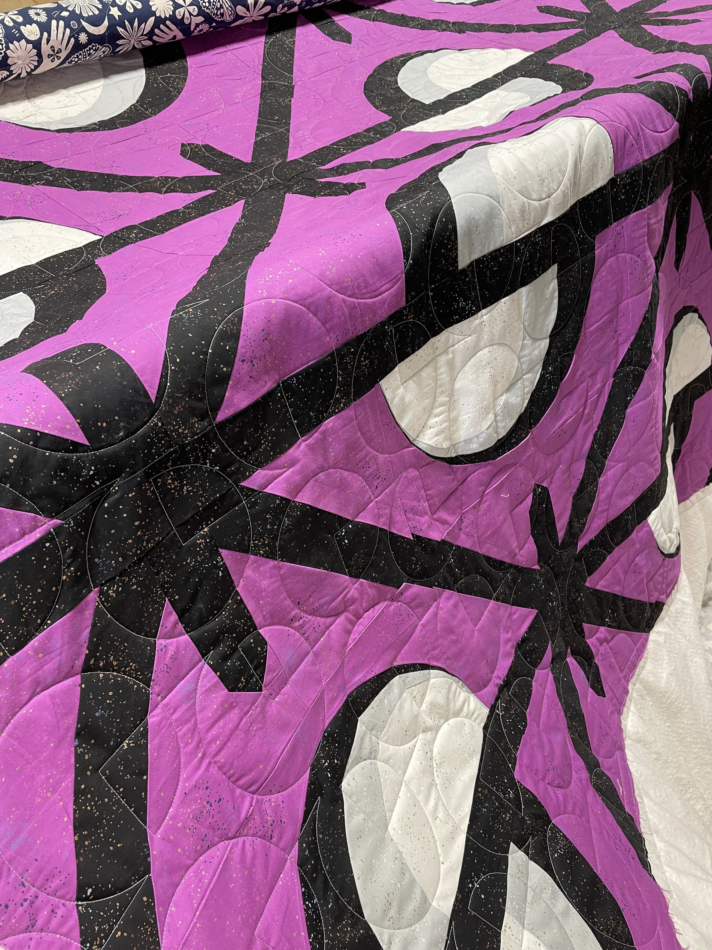 A quilt with a purple background featuring a black spider web pattern and white circular accents, with a black spider motif.