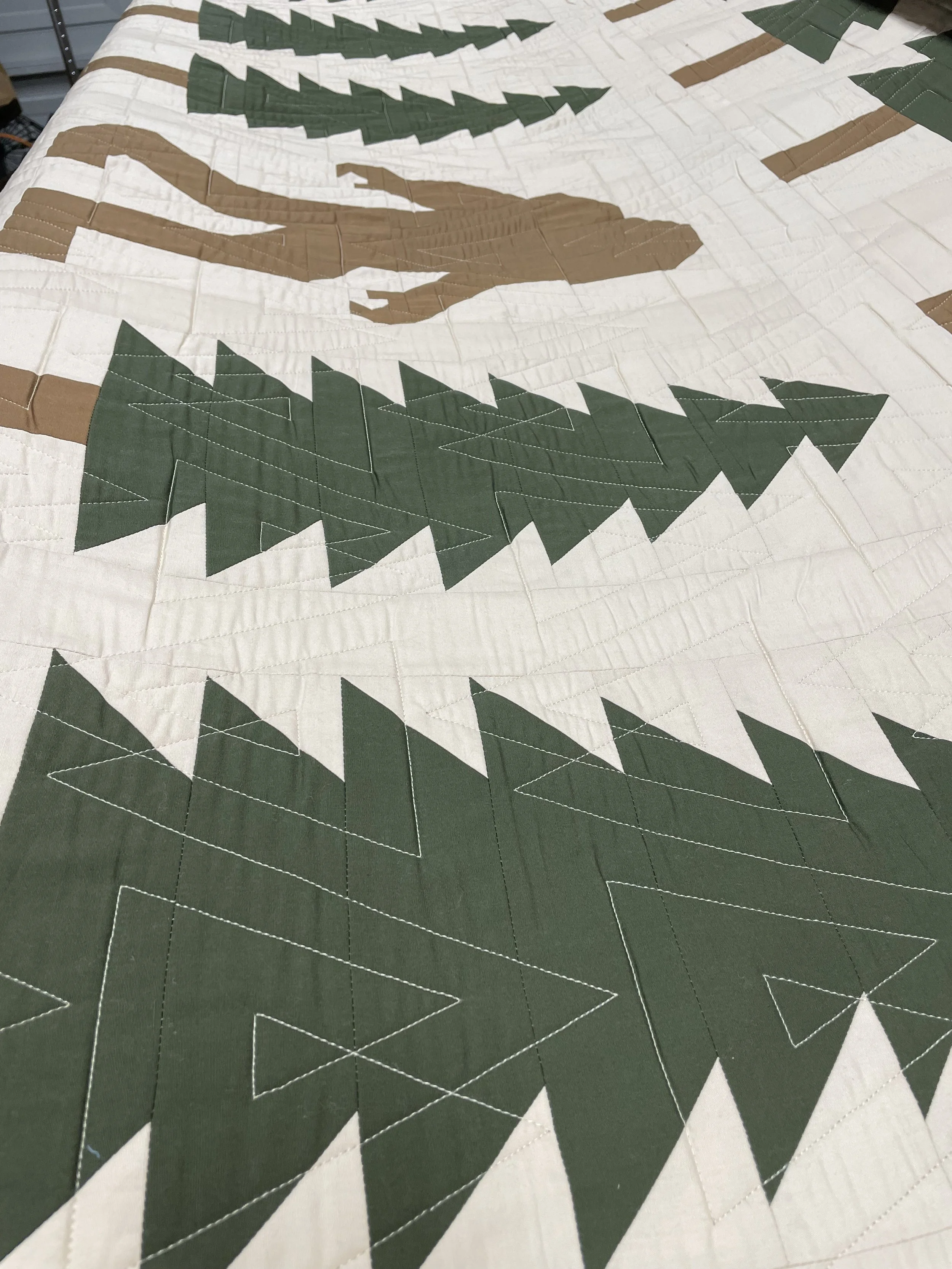 Close-up of a quilt with a mountain and trees design in green, brown, and white colors, featuring triangular shapes and detailed stitching.