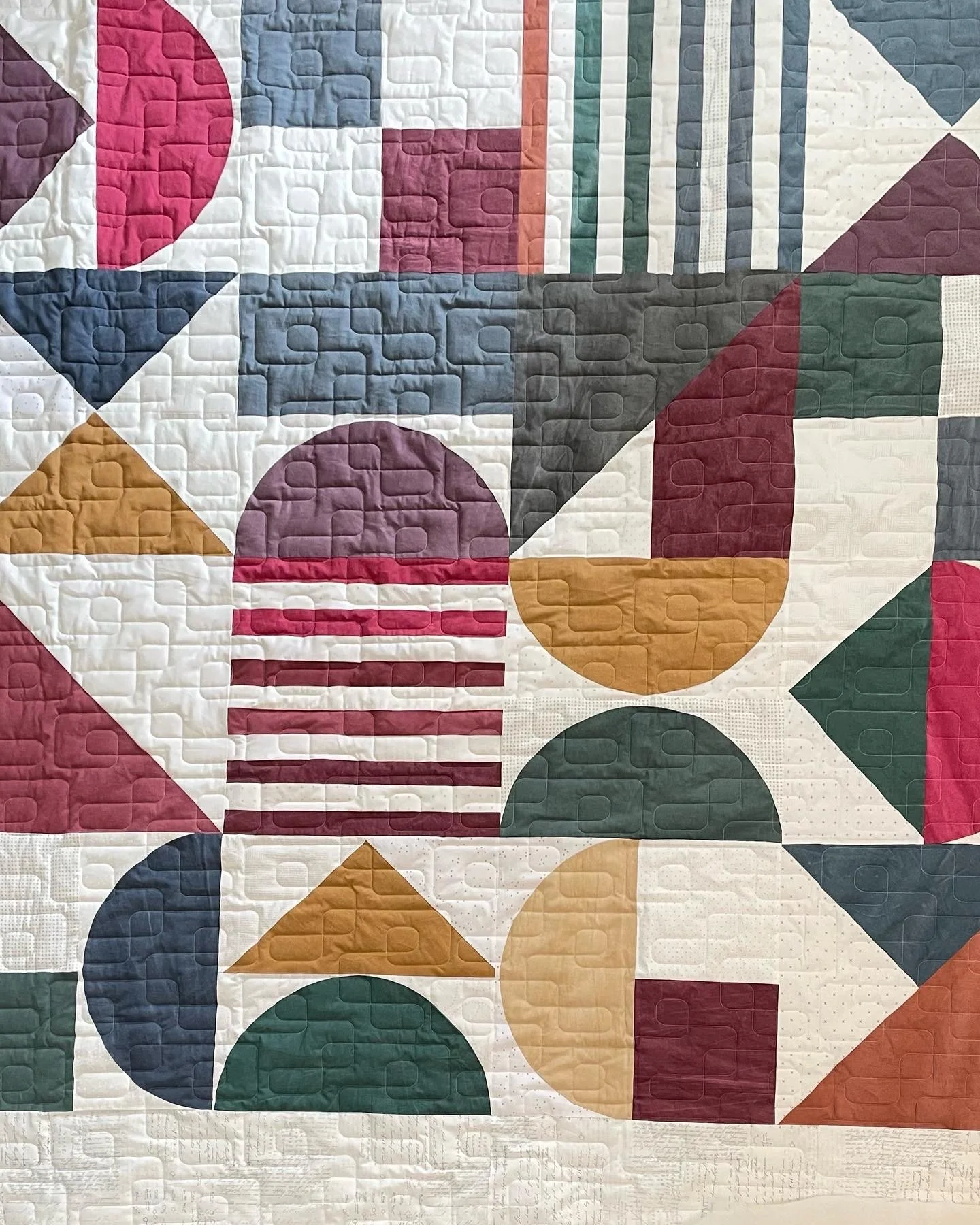 Colorful geometric quilt with various patterns including circles, triangles, and stripes in colors like pink, red, navy, ochre, green, dark grey, and white.