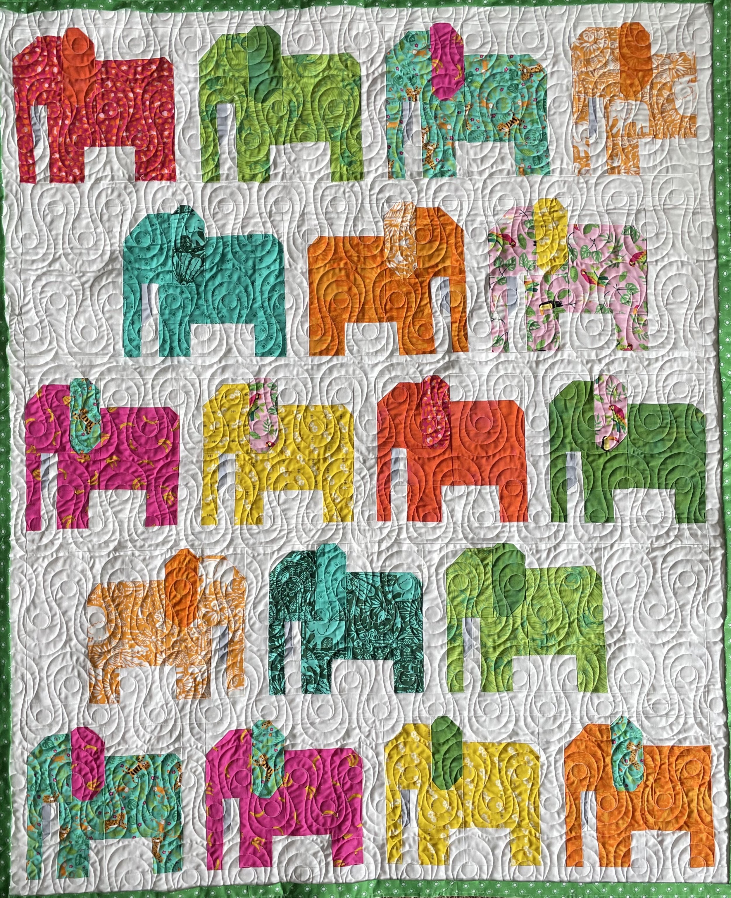 A colorful quilt with a pattern of various elephants in pink, green, orange, turquoise, yellow, and floral prints, separated by white background with intricate quilting, bordered with a green polka dot fabric.