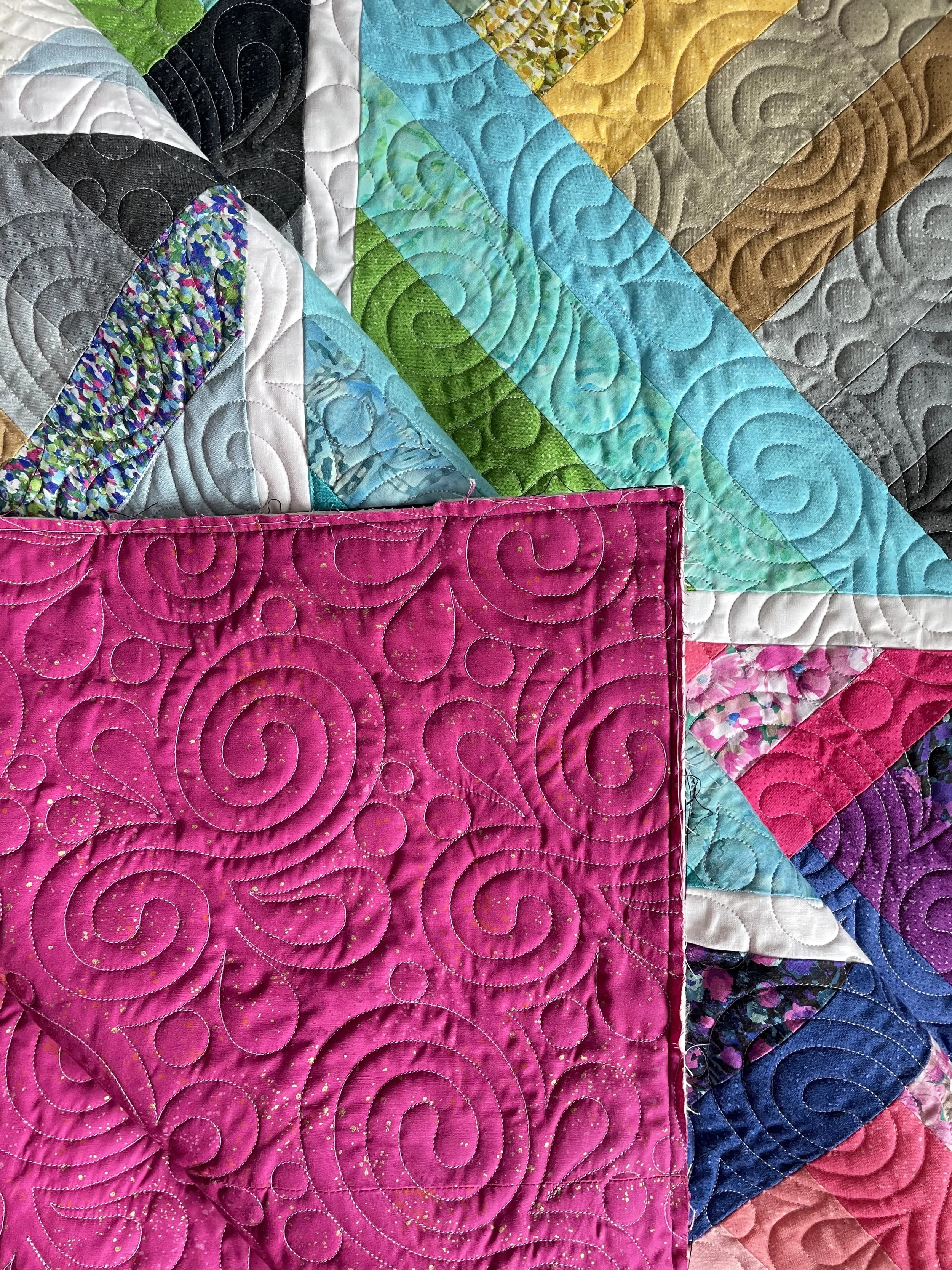 Close-up of a colorful quilt with various patterned fabric pieces and detailed stitching, featuring a pink section with swirling quilted design.