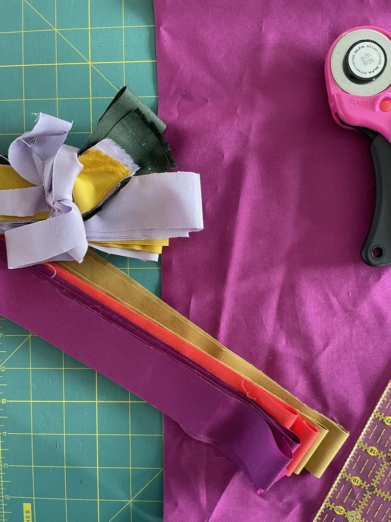 Sewing or crafting workspace with colorful fabric, including purple and pink, along with strips of metallic gold and red fabric, a rotary cutter, and a green cutting mat with yellow measurement lines.