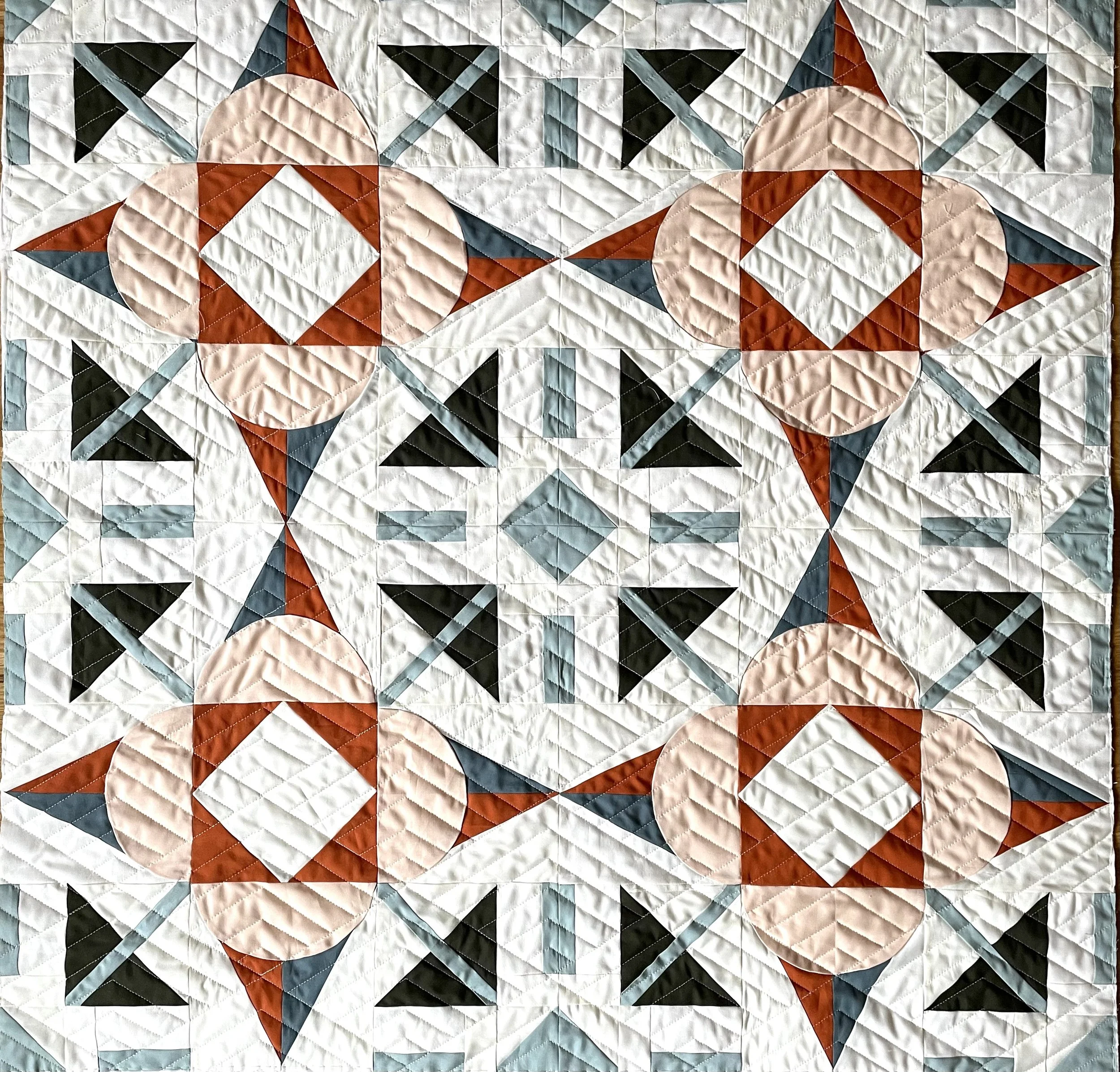 A geometric quilt design with a pattern of star shapes, circles, and squares in colors including white, black, rust, peach, and teal.