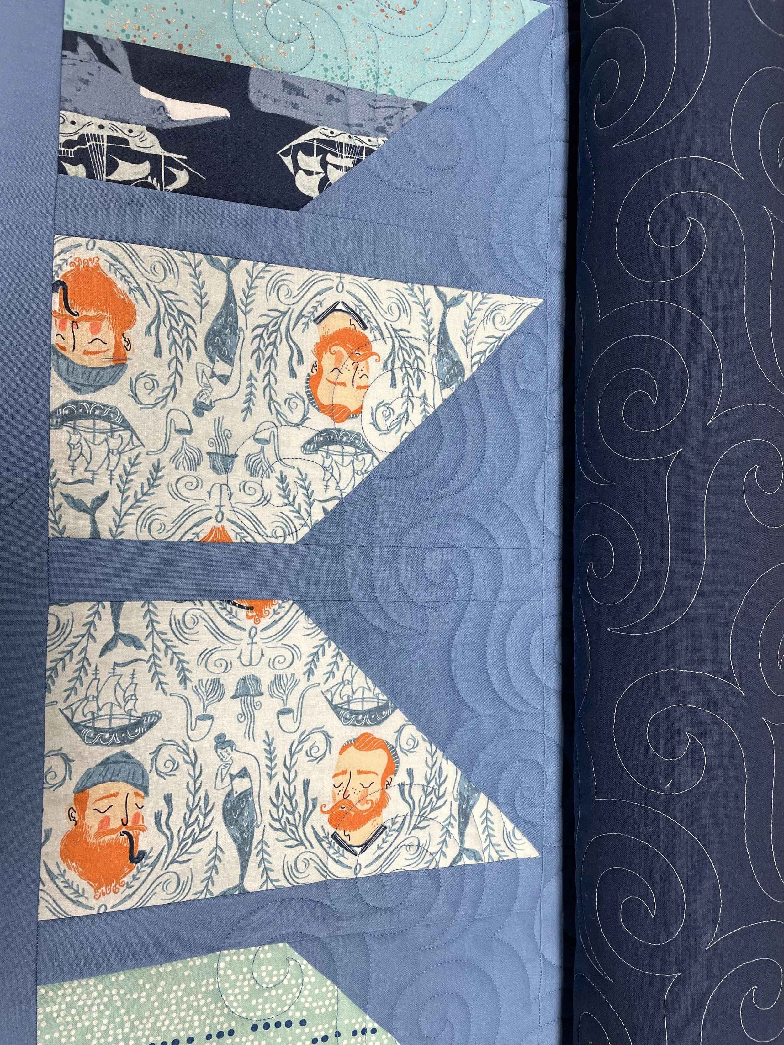 Close-up of a quilt with nautical and maritime-themed fabric triangles and a blue quilted border with swirl patterns.
