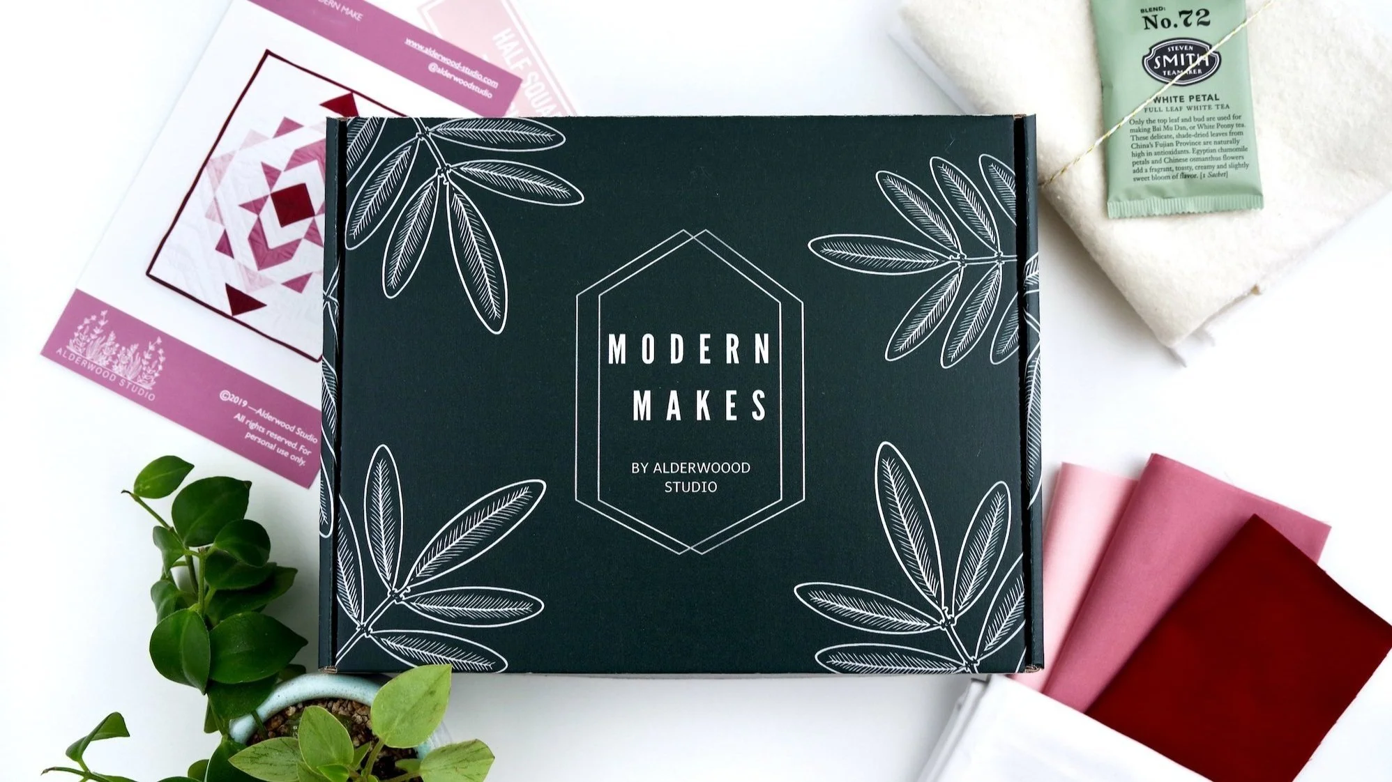 Flat lay of crafting materials including a black box labeled 'Modern Makes by Alderwood Studio' with white leaf illustrations, pink, red, and white fabric swatches, a white towel with a green tea bag on top, printed instructions or pattern sheet, and a small green potted plant on a white background.