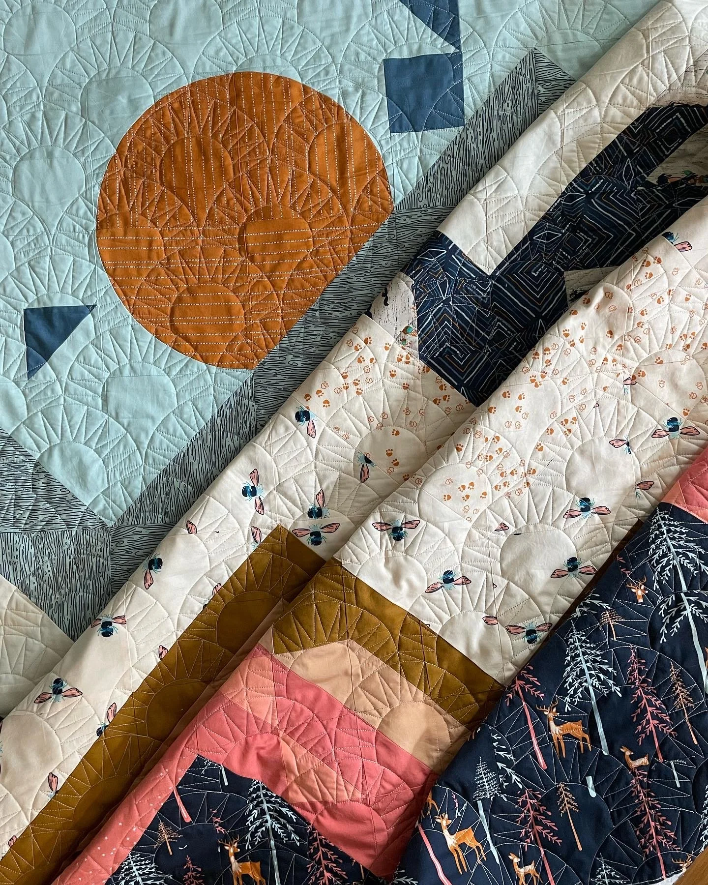 Close-up of colorful patchwork quilts with various patterns including geometric shapes, bees, trees, and animals, in shades of blue, pink, cream, and orange.