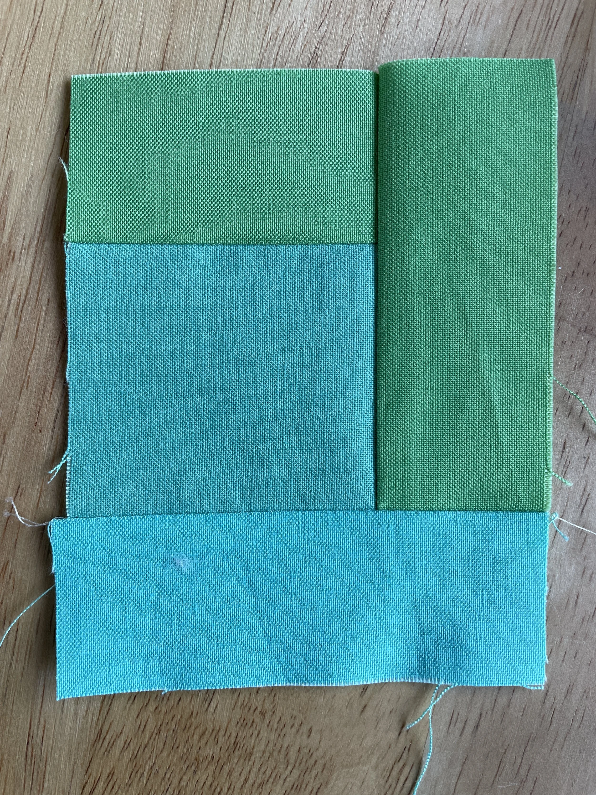 Four pieces of fabric in shades of green and blue sewn together in a block pattern on a wooden surface.