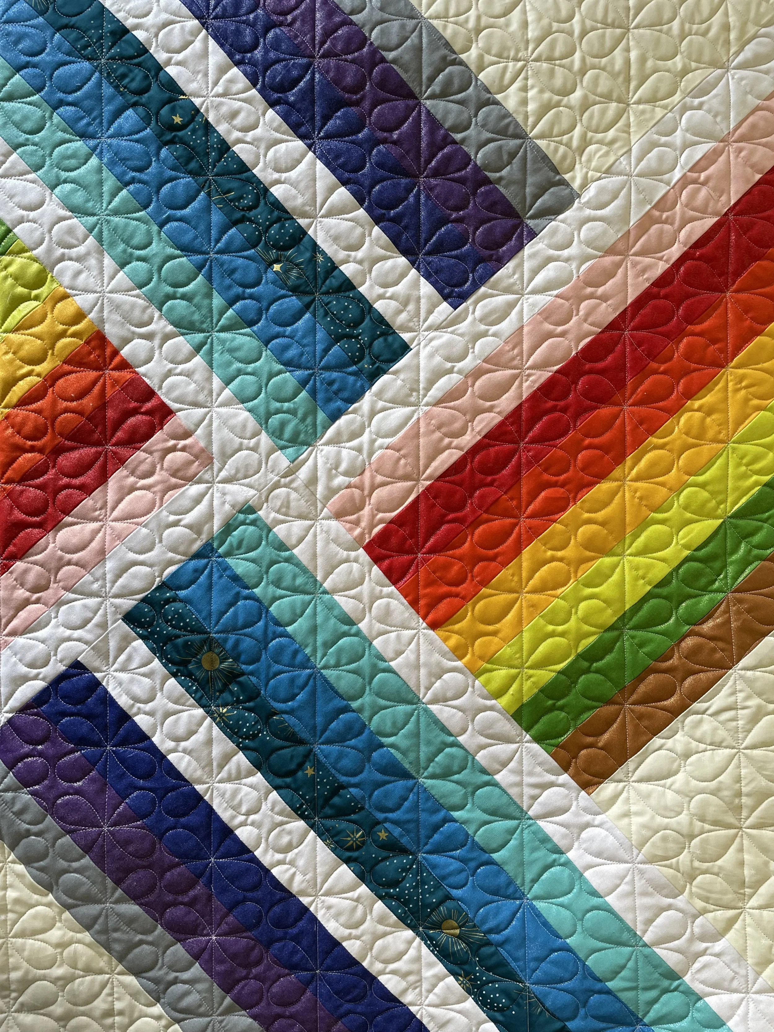 Close-up of a colorful quilt with diagonal stripes in various colors like blue, green, red, yellow, purple, and white, featuring a detailed quilted pattern of intersecting loops.