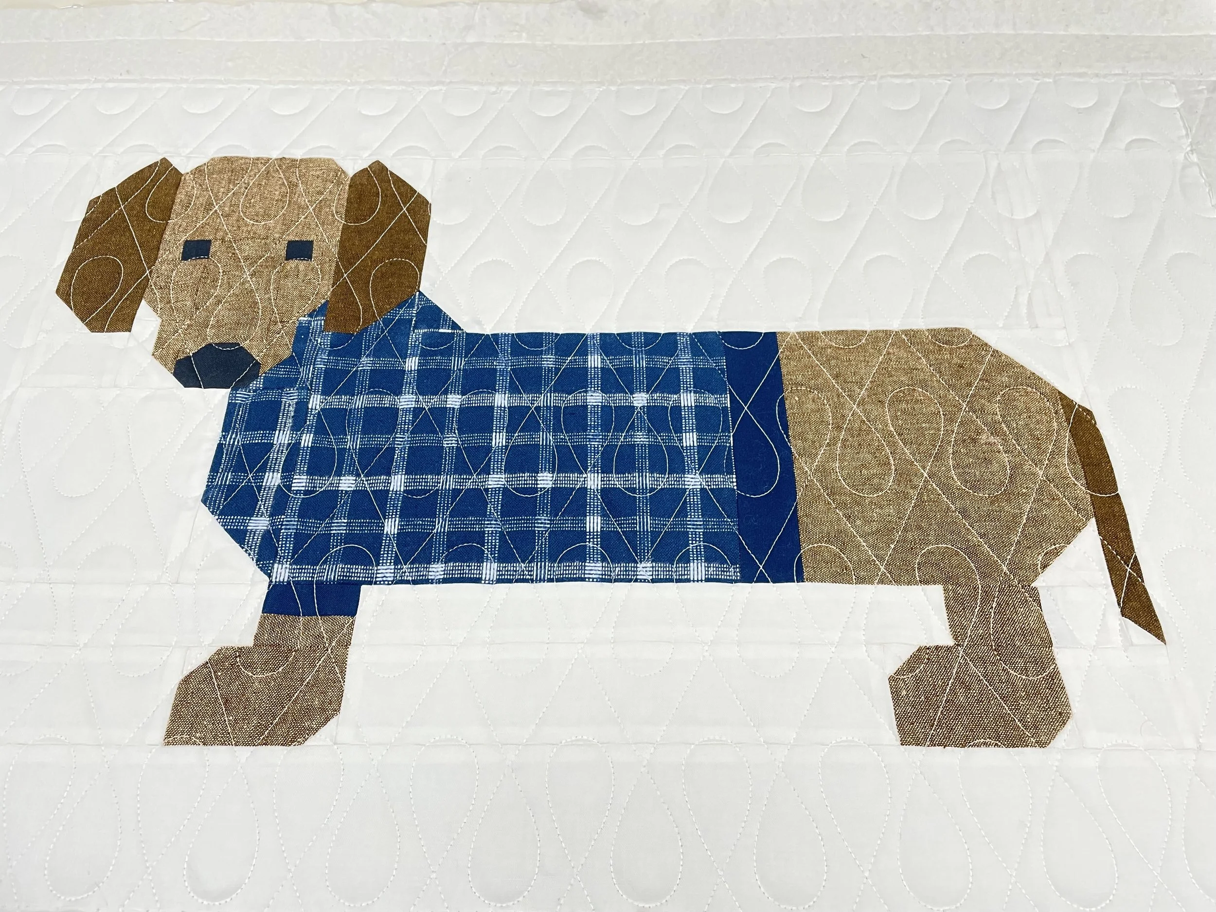A quilted fabric artwork of a dog, with a brown head, tan ears and tail, wearing a blue checkered shirt, on a white background.