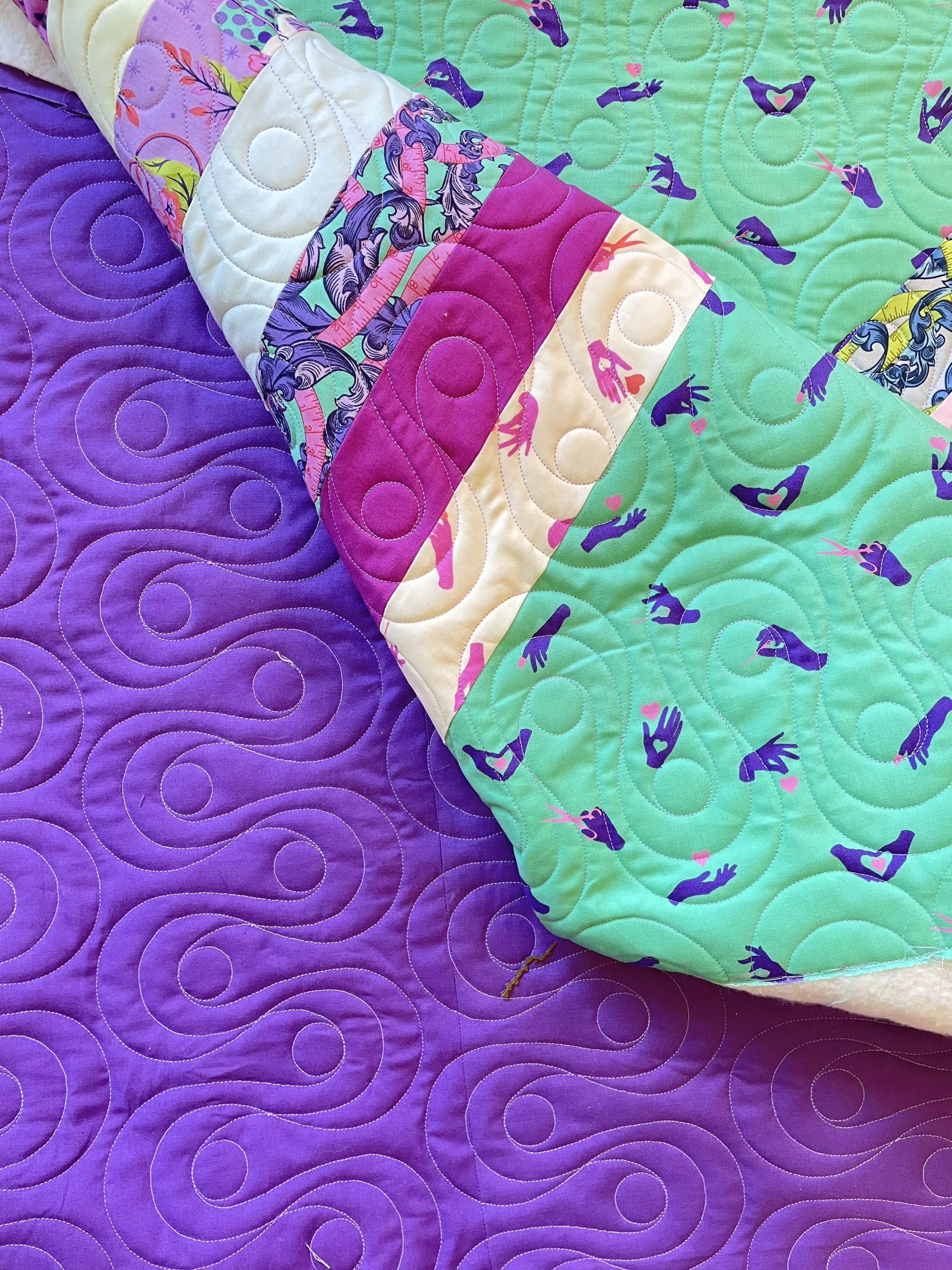 Colorful quilting with a purple background and bright, patterned quilted pieces featuring hands making heart and love symbols, and mermaids, arranged in a layered fashion.