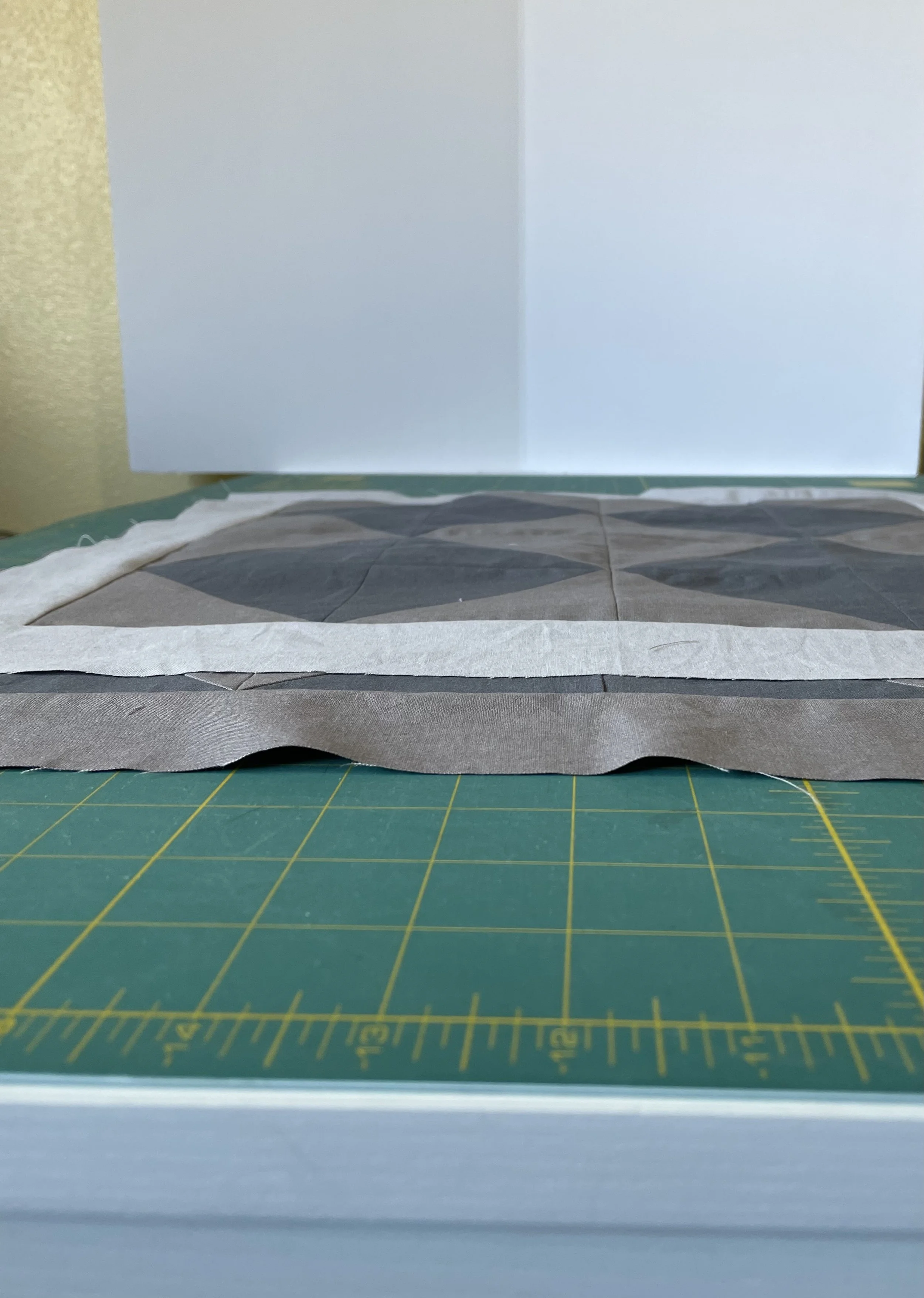 A scalloped-edge fabric with a geometric pattern in shades of gray, black, and white, placed on a green cutting mat with a yellow grid, with a plain white background.