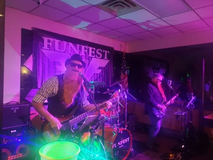 Funfest, with colorful stage lights. The band includes a bassist in sunglasses and a checkered shirt, a drummer in the background, and a guitarist. An American flag is visible near the stage.