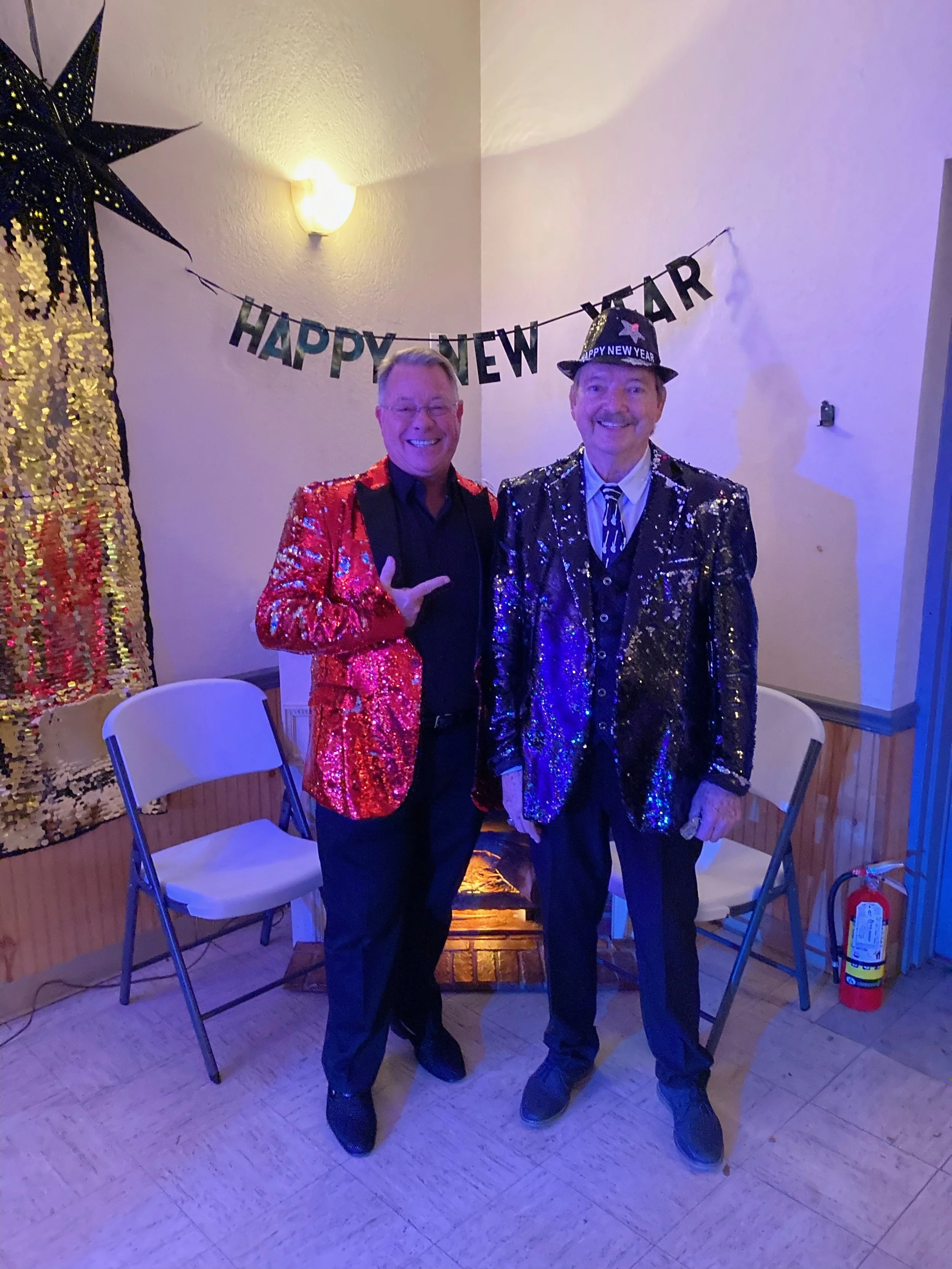 Two men in glittering red and black jackets celebrating New Year's, standing in front of a "HAPPY NEW YEAR" banner, with festive decorations including a large star-shaped lantern and sequin backdrop.