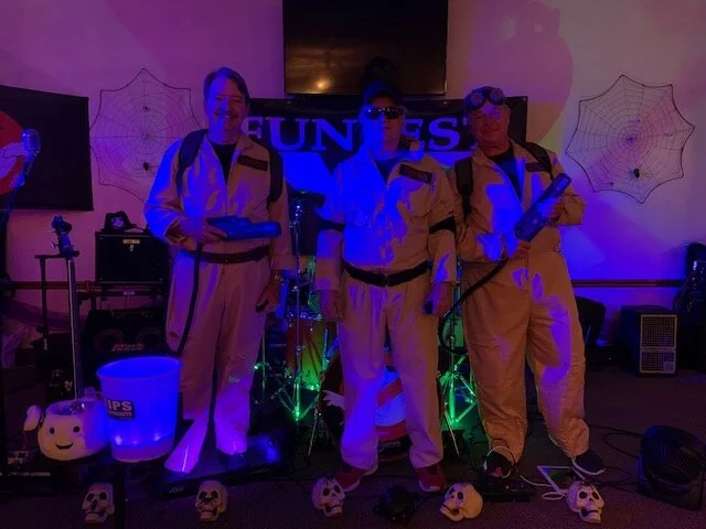 FunFest Band dressed as ghostbusters in costumes standing in front of a stage with spooky decorations, including spider webs, with musical equipment behind them and colorful lighting.