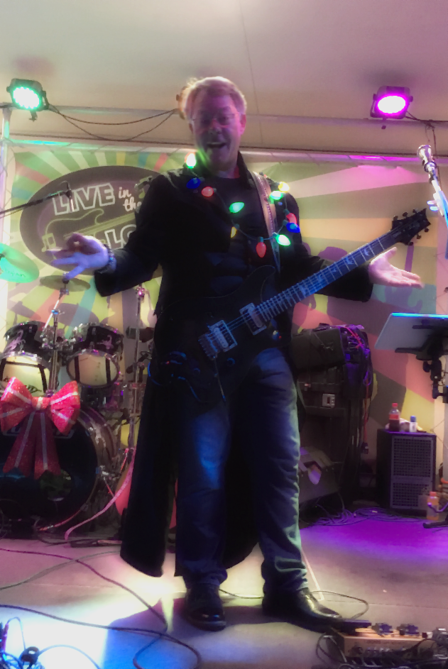 Holt performing on stage with an electric guitar, decorated with colorful string lights, in front of a drum set with a large red bow. The stage features bright, multicolored lights and a backdrop with musical-themed text.