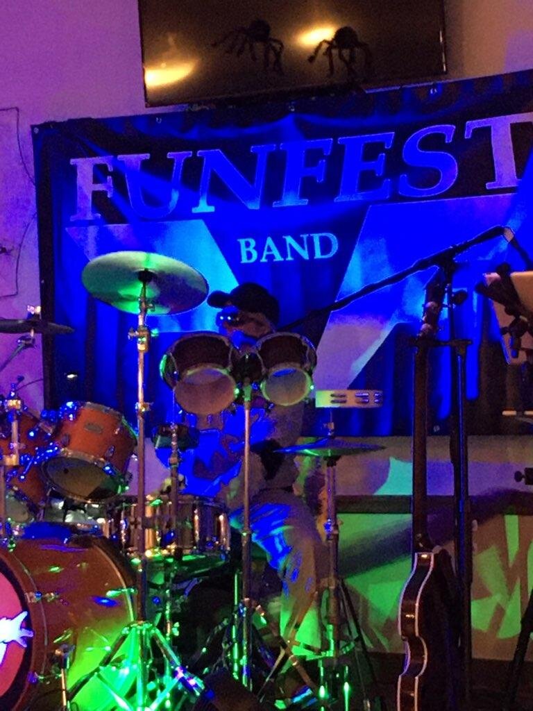 A band performing on stage with drums and a person playing a percussion instrument, behind a banner that says 'FUNFEST BAND'. Colored stage lights create a vibrant atmosphere.