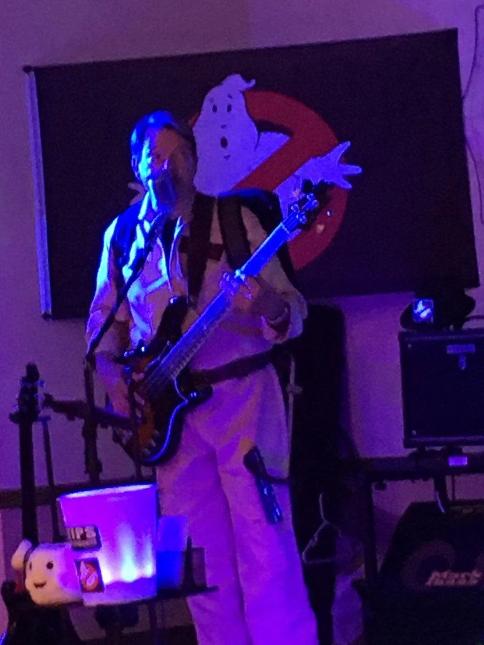 Person playing guitar on stage, wearing glasses and a burglary costume, with a Ghostbusters banner in the background.