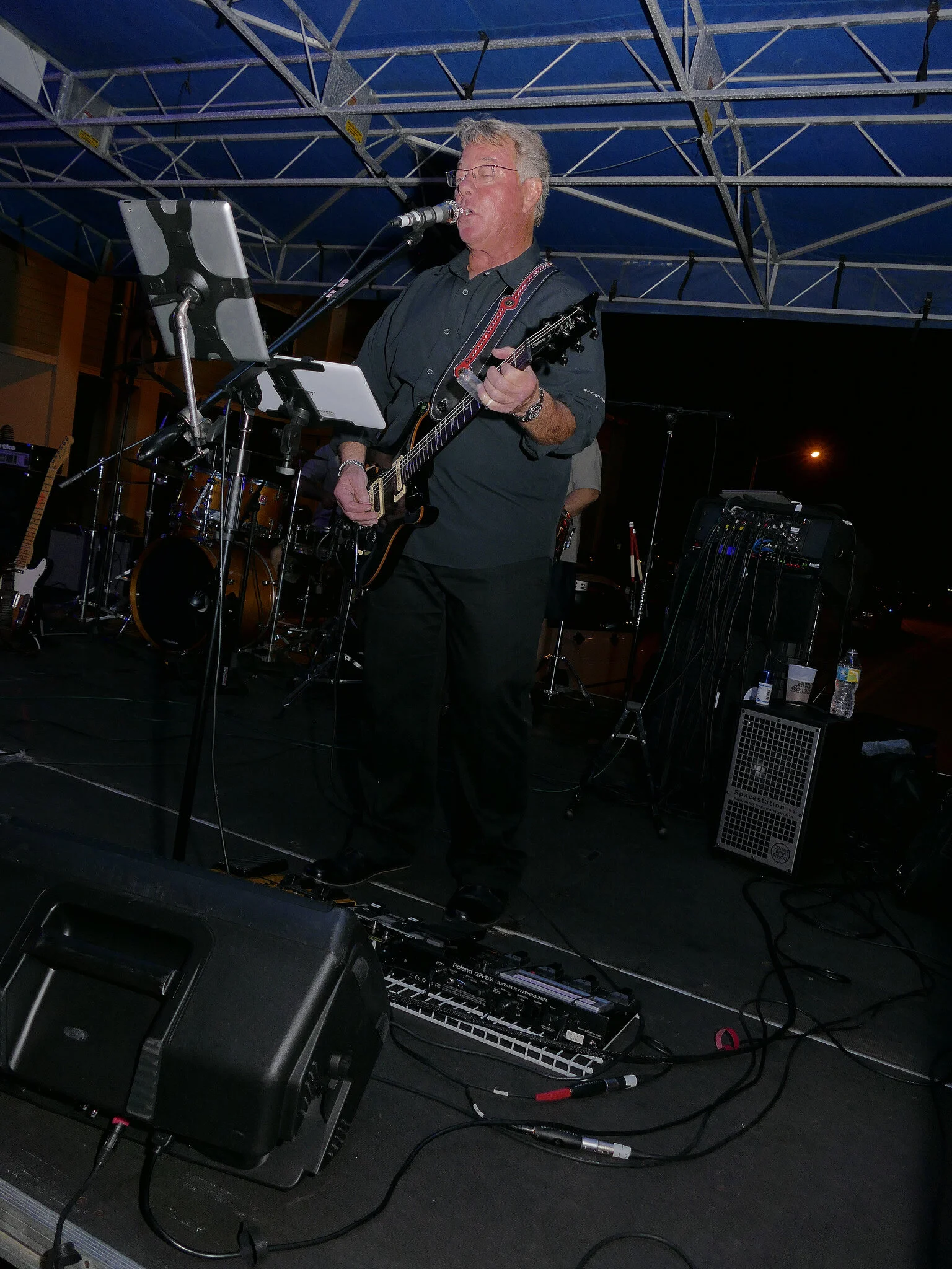 A man with glasses in a black shirt playing an electric guitar and singing into a microphone during a live music performance at night under a metal canopy.
