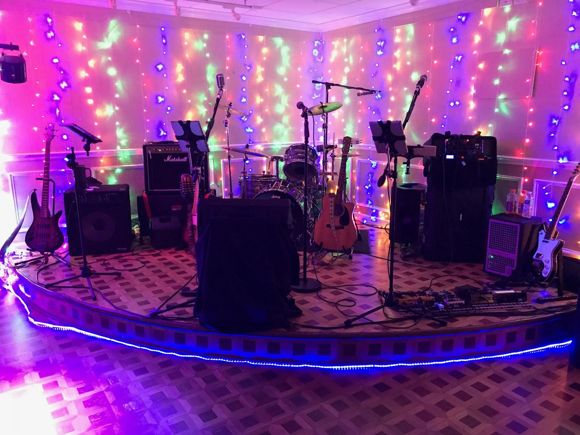A music stage setup with various instruments including guitars, drums, and microphones, decorated with colorful LED lights at a performance venue.
