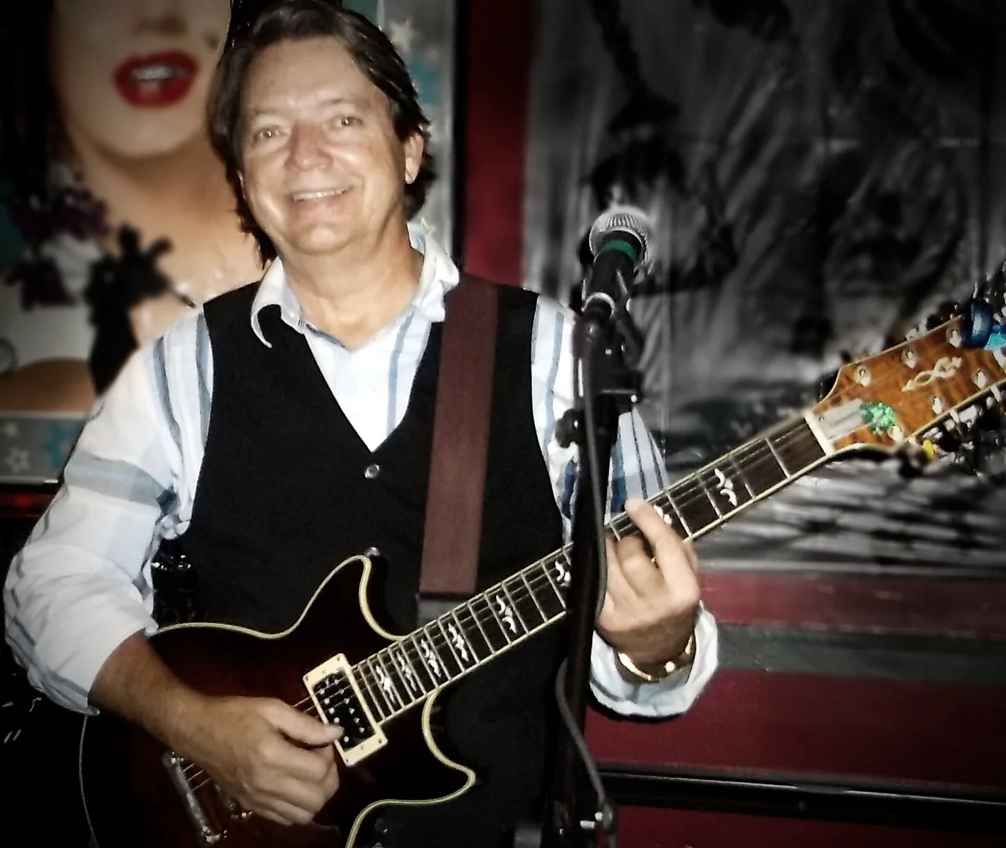 A man playing an electric guitar and smiling, standing in front of a microphone with a large poster of a woman in the background.