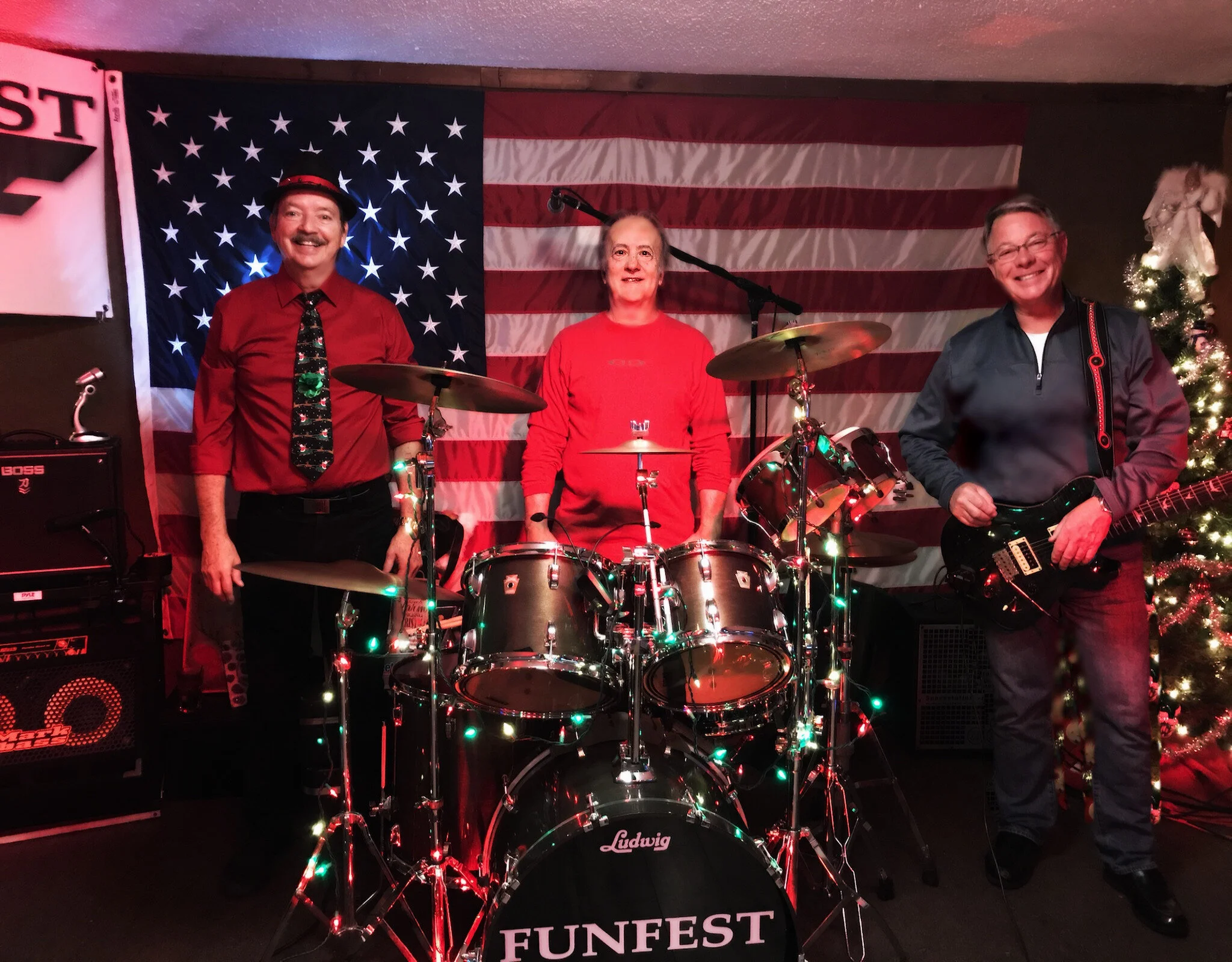 Three people in front of a large American flag and Christmas tree, standing around a drum set decorated with Christmas lights. The person on the left is wearing a red shirt and tie, the person in the middle is wearing a red shirt, and the person on t