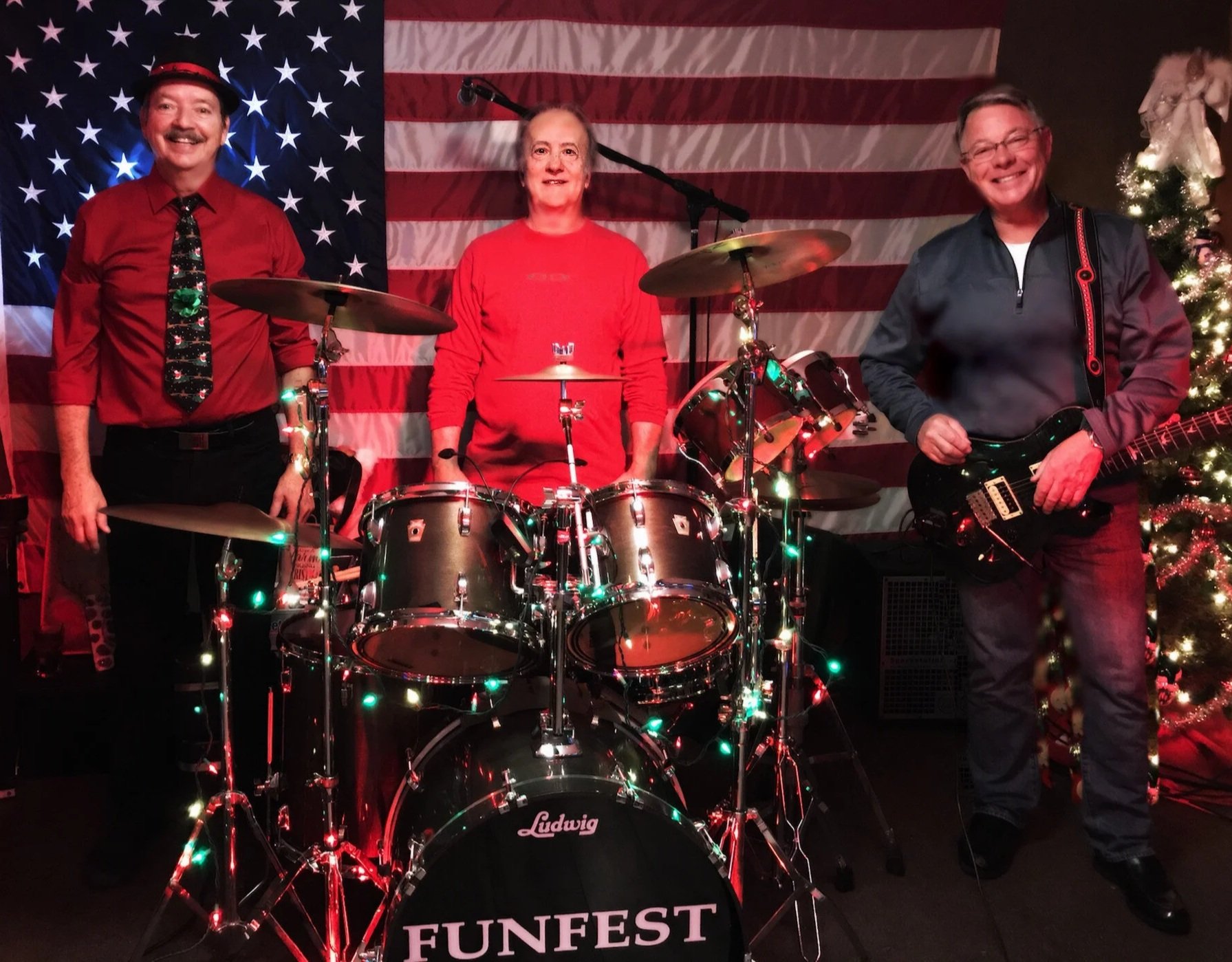 Three people in front of a large American flag and Christmas tree, standing around a drum set decorated with Christmas lights. The person on the left is wearing a red shirt and tie, the person in the middle is wearing a red shirt, and the person on t