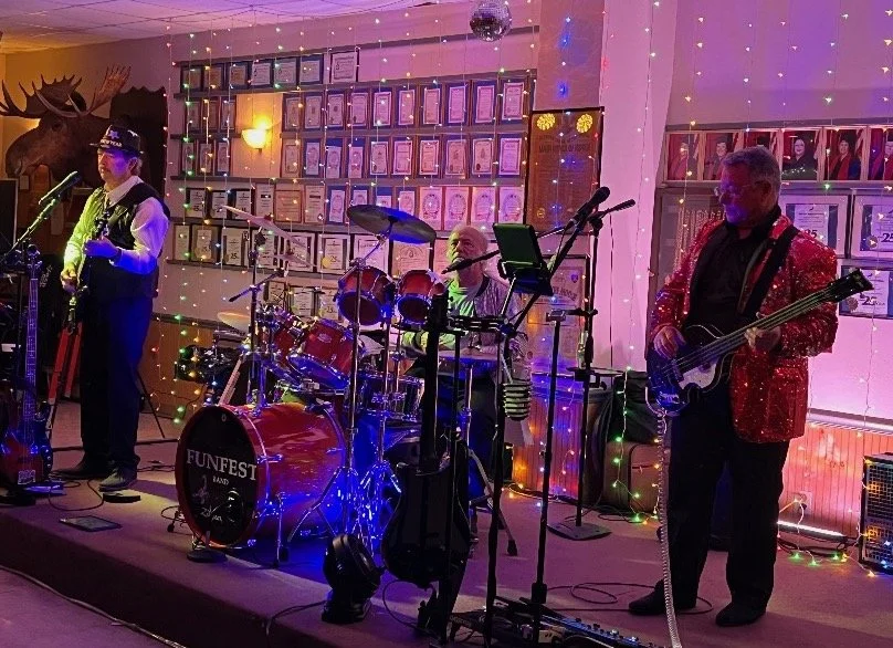 Band performing live on stage with colorful lights in a decorated venue, including framed pictures on the wall and a mounted moose head.