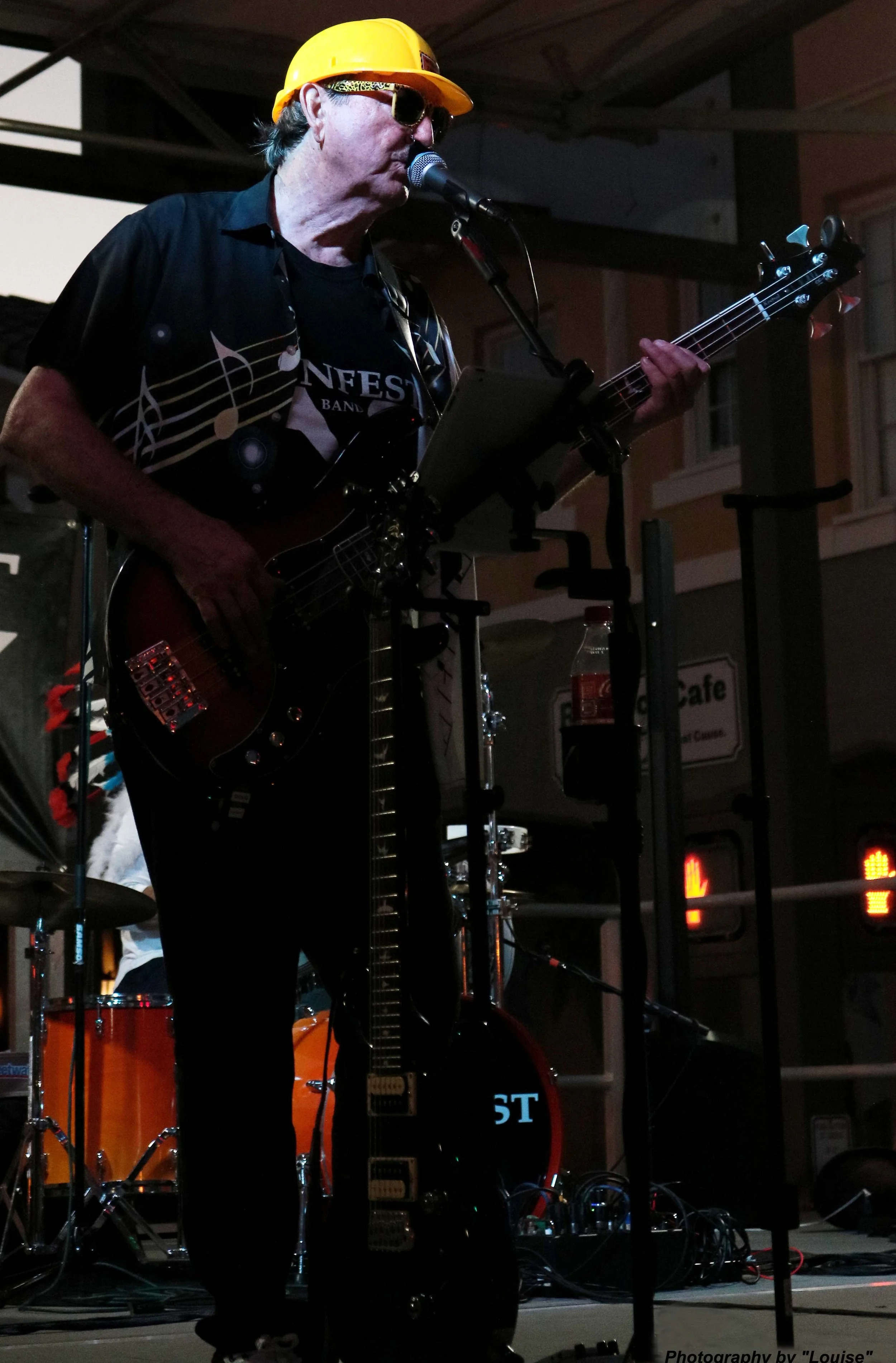 A musician wearing sunglasses and a yellow hard hat is playing an electric bass guitar and singing into a microphone on stage during a performance.