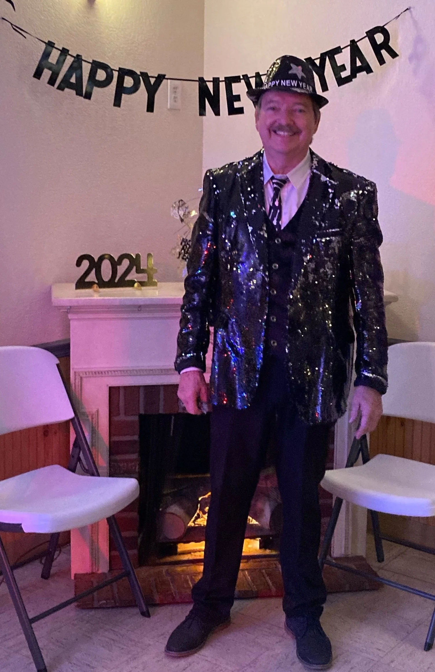 A man dressed in a shiny, black, sequin-covered jacket, black pants, a striped tie, and a Darth Vader-themed party hat, standing in front of a fireplace decorated with a '2024' sign and a 'Happy New Year' banner hanging on the wall, celebrating New Y
