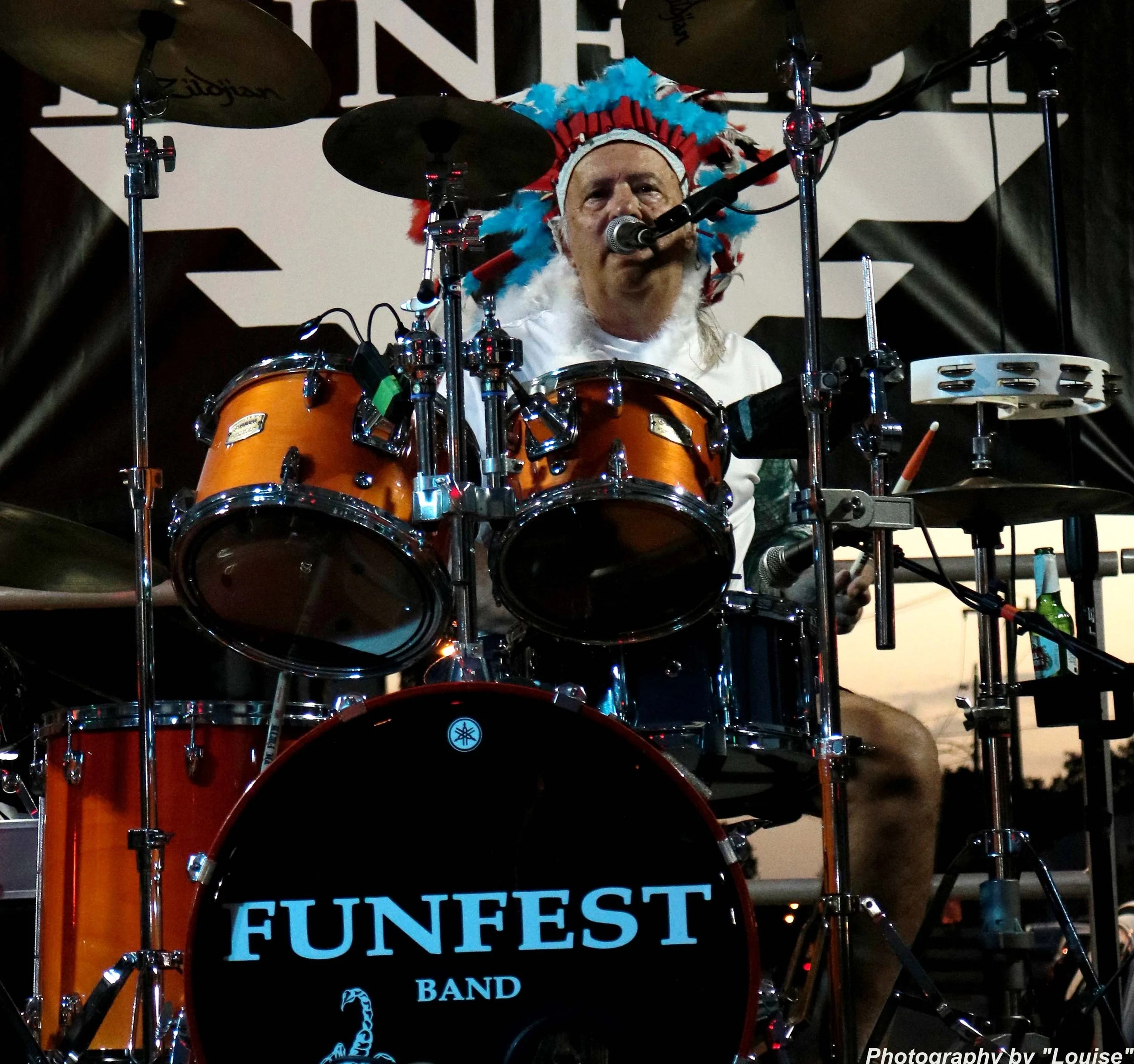 A person dressed as a Native American chief playing a drum set on stage at a band event called FUNFEST, with a cloudy sky in the background.