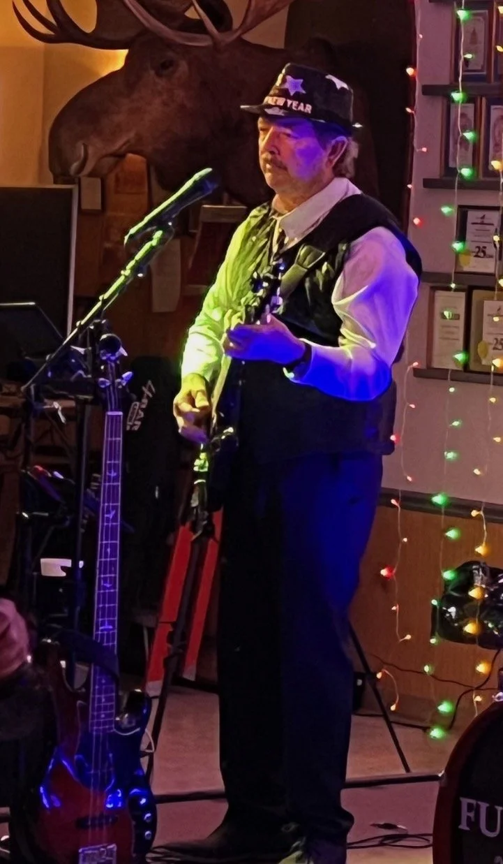 Man wearing a birthday hat and vest, playing guitar and singing into a microphone in a decorated, dimly lit venue with a mounted moose head in the background.