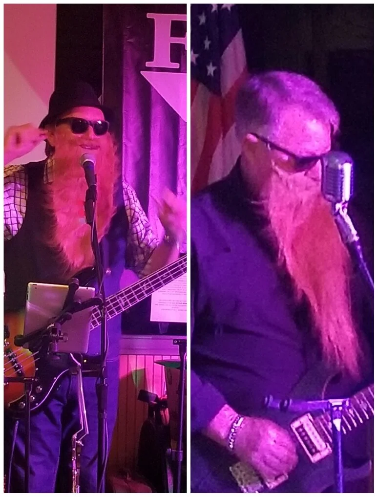 Two musicians performing on stage. The musician on the left has a long, red beard, sunglasses, wearing a black hat, and playing guitar, with a microphone and tablet in front. The musician on the right has long, red beard, glasses, wearing dark clothi