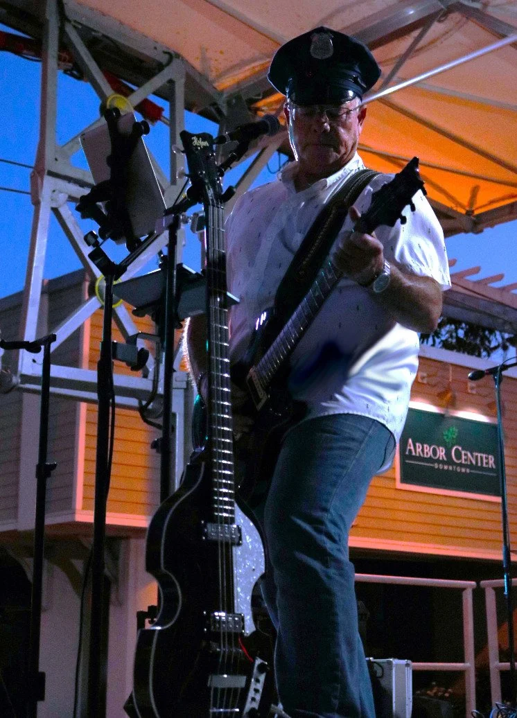 A man wearing a police hat, glasses, and a white shirt is playing an electric guitar on an outdoor stage at dusk. There are microphones and music equipment around him, and a sign that reads 'Arbor Center' is visible in the background.