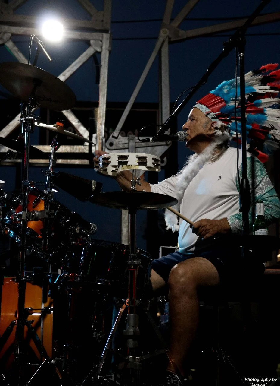 A man wearing a Native American headdress is playing drums on a stage at night.