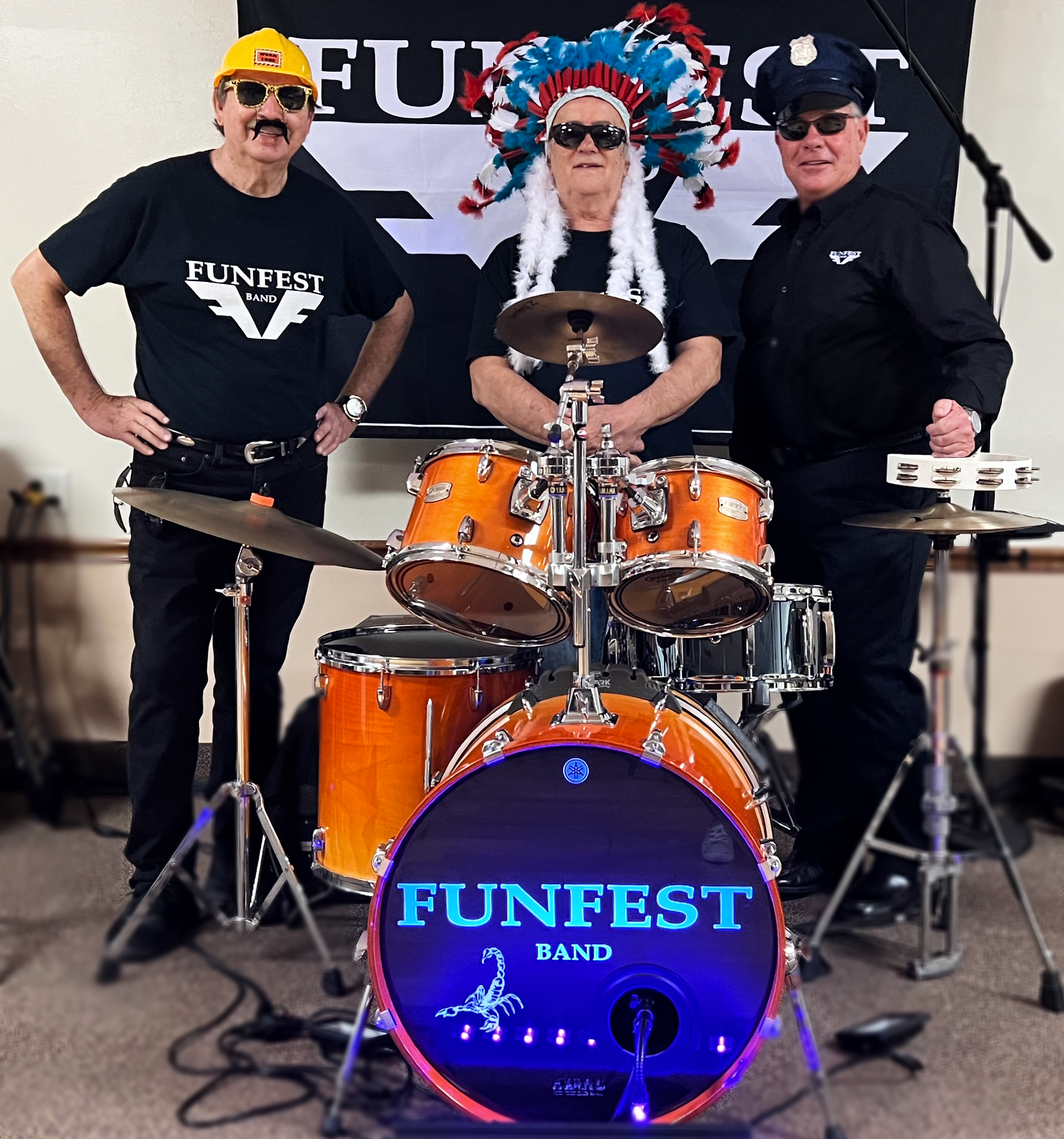 Three band members of FUNFEST BAND posing behind a drum set with a glowing bass drum. The woman in the middle is wearing a colorful feather headdress. The man on the left is wearing sunglasses and a yellow hard hat, and the man on the right is dresse