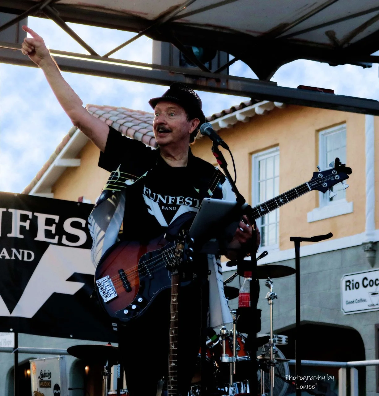 A man playing bass guitar on an outdoor stage, pointing with his right hand, wearing a black hat and a black t-shirt with music notes and the words 'Unfest Band' on it, with a microphone in front of him and a drum set behind him.