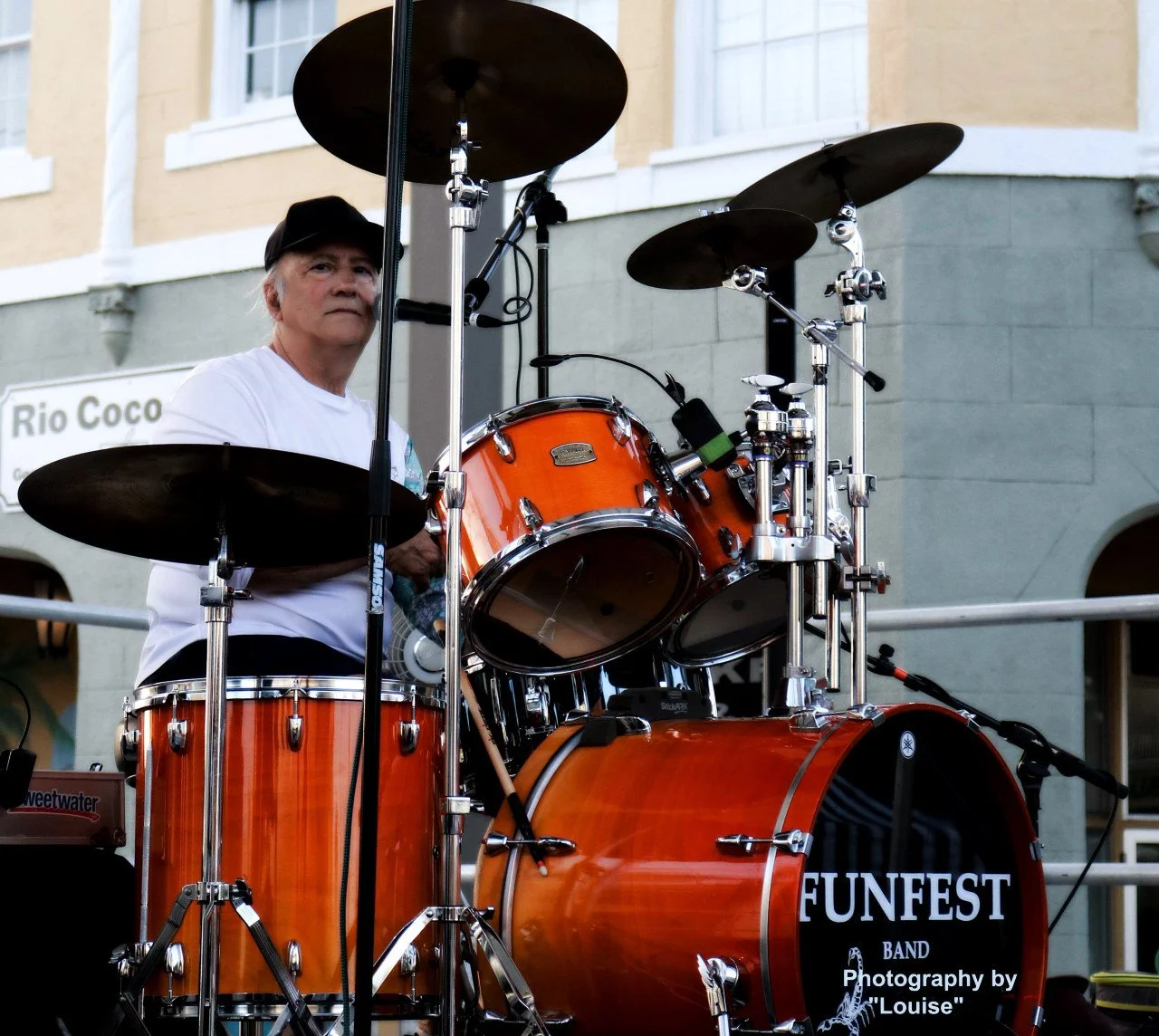 An older man playing an orange drum set during a public event, with a sign that reads 'FUNFEST BAND' and a photo credit to 'Louise' in the lower right corner.