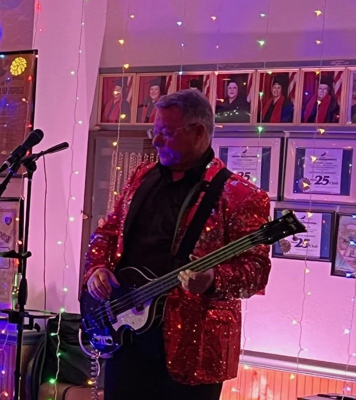 A man playing bass guitar in a decorated room with framed photos and awards on the wall, colorful string lights, and a microphone stand nearby, wearing a sparkly red jacket and glasses.