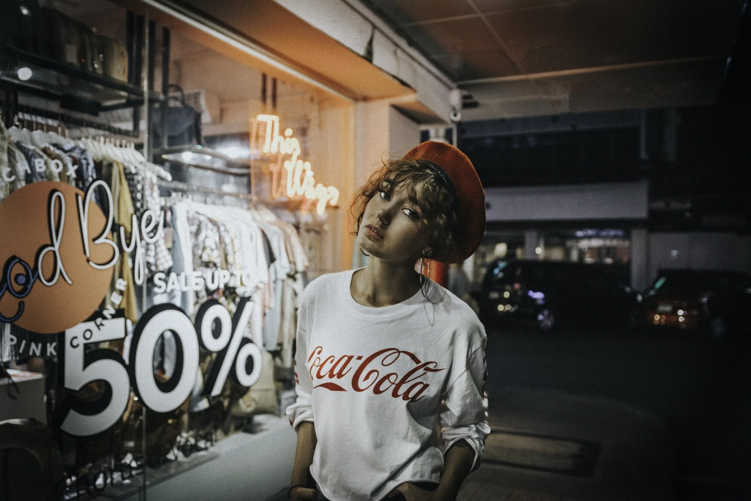 A woman in a coca-cola white sweatshirt stands in front of a 50% sign in a store.
