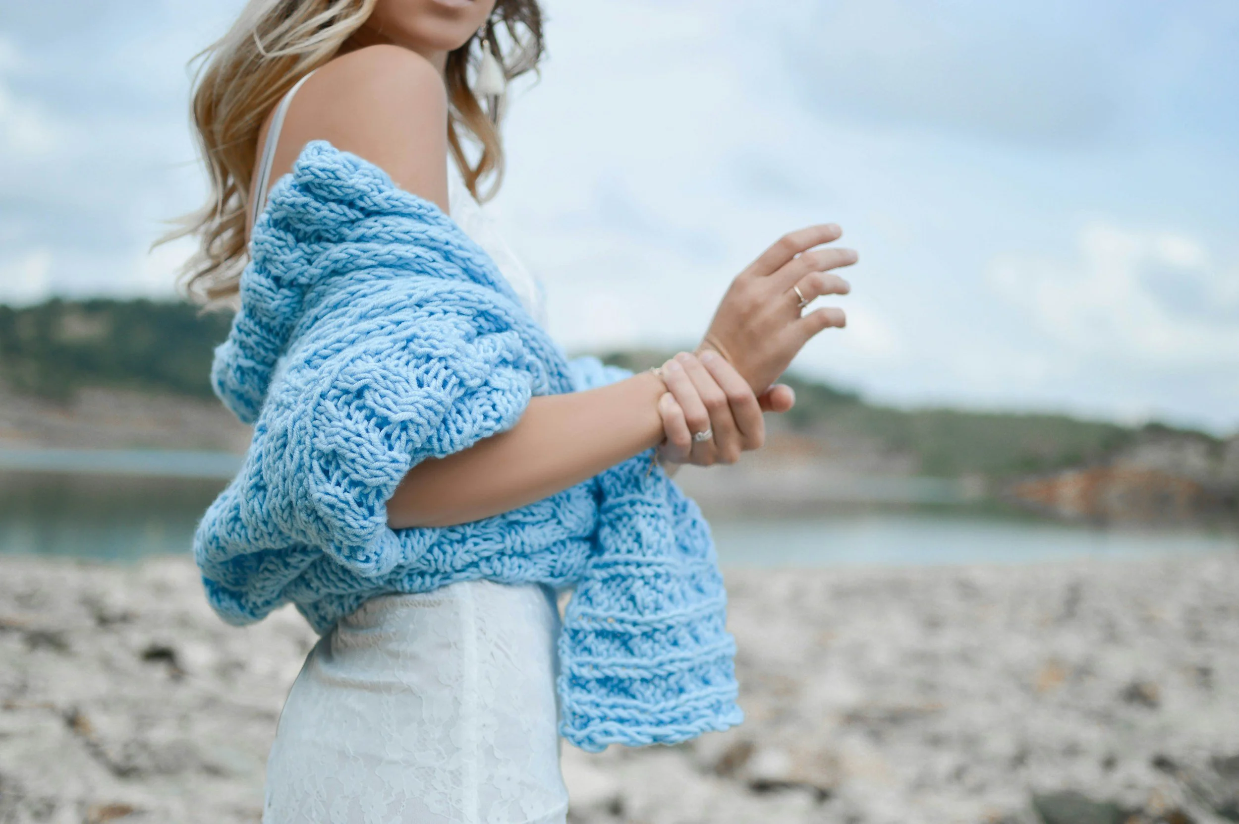 A blonde woman stands on a beach while wearing a blue knit cardigan.