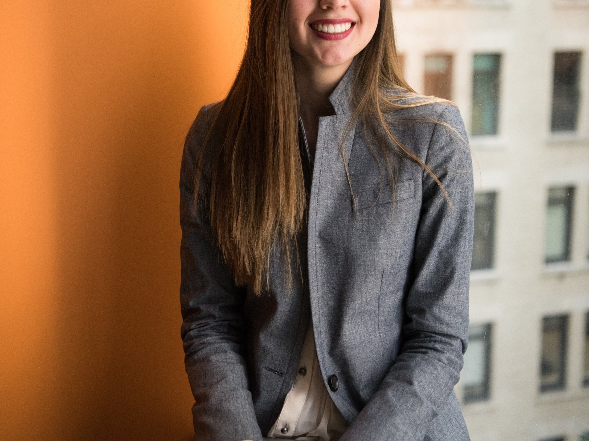Woman with long brown hair stands against an orange wall wearing a grey blazer.